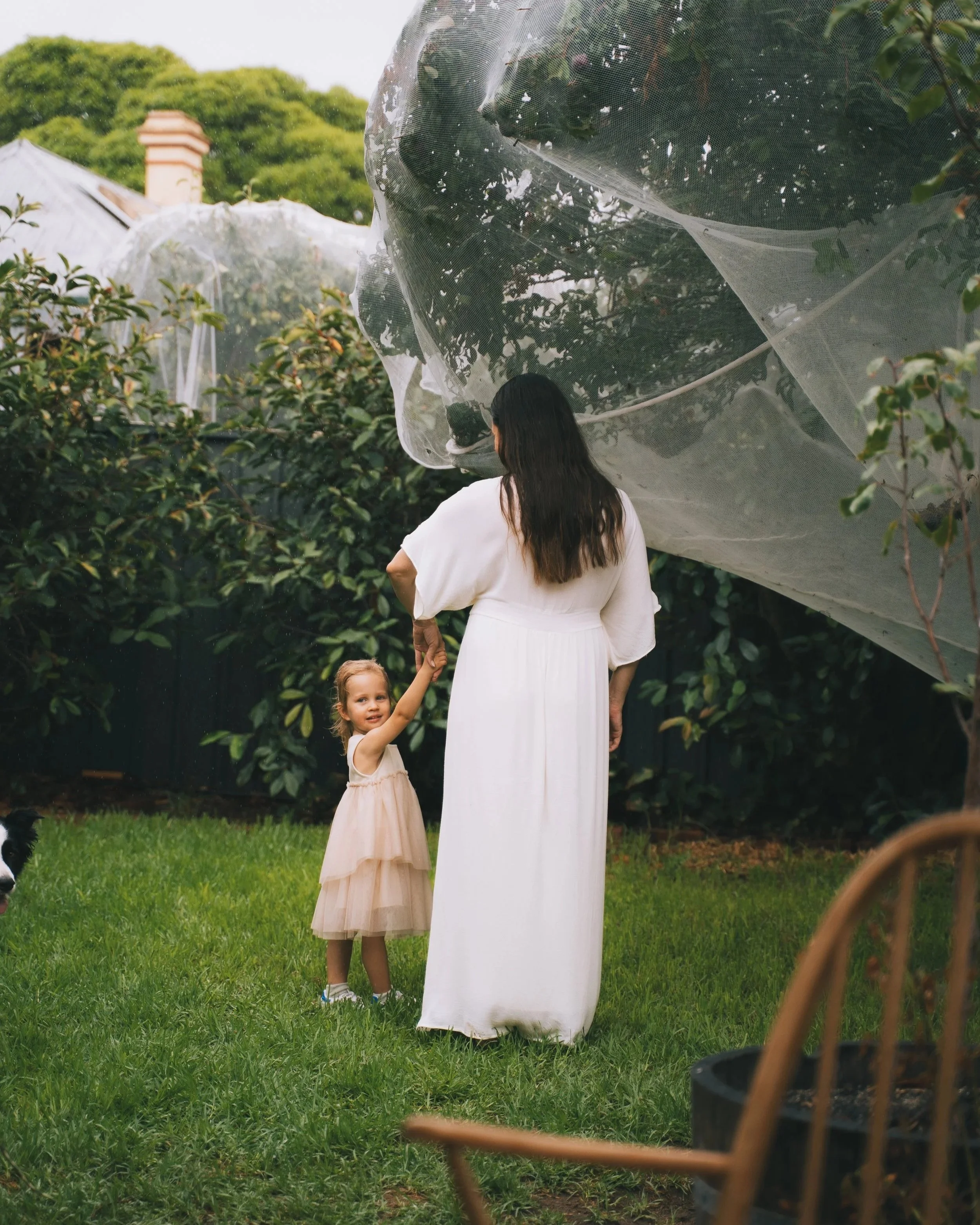 A woman in a white dress holding hands with a little girl in a light pink dress outside in a garden, with trees and greenery in the background.