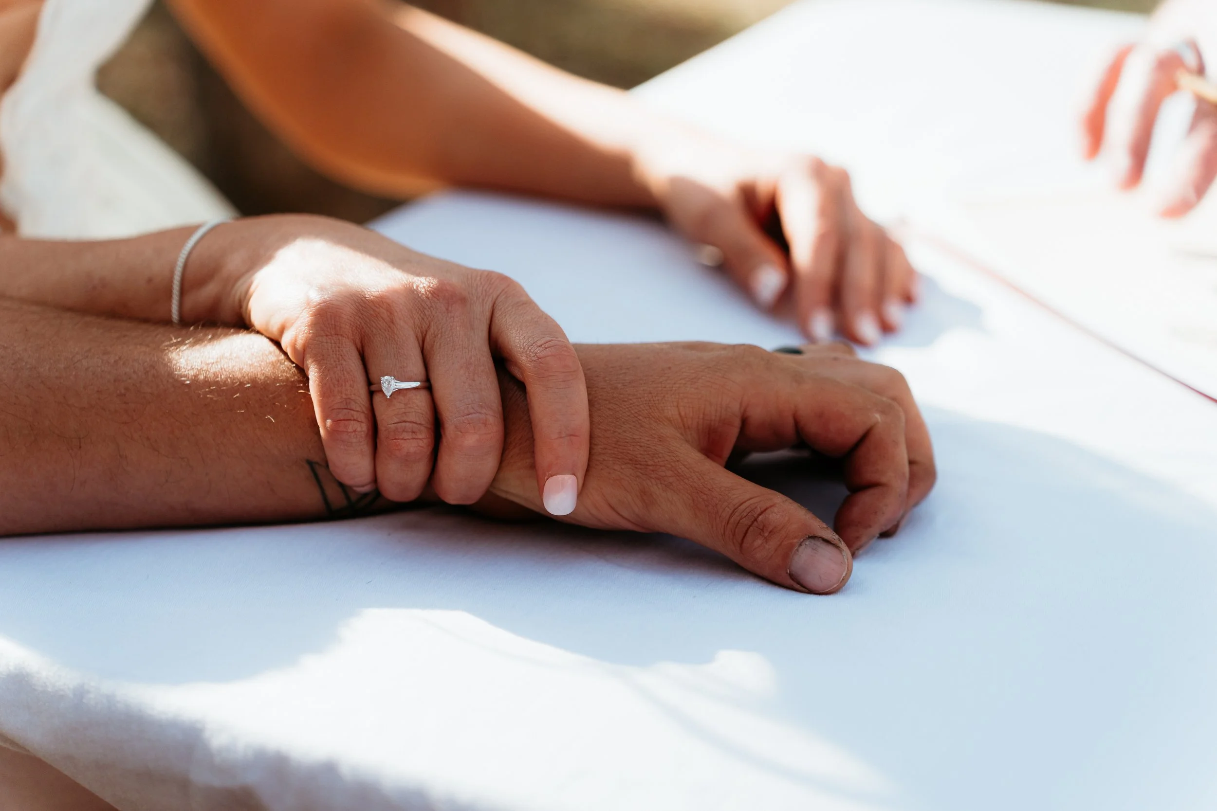 Close-up of a newlywed couple's hands, with the bride's hand resting on her groom's hand, showing an engagement ring and wedding band, outdoors with sunlight.