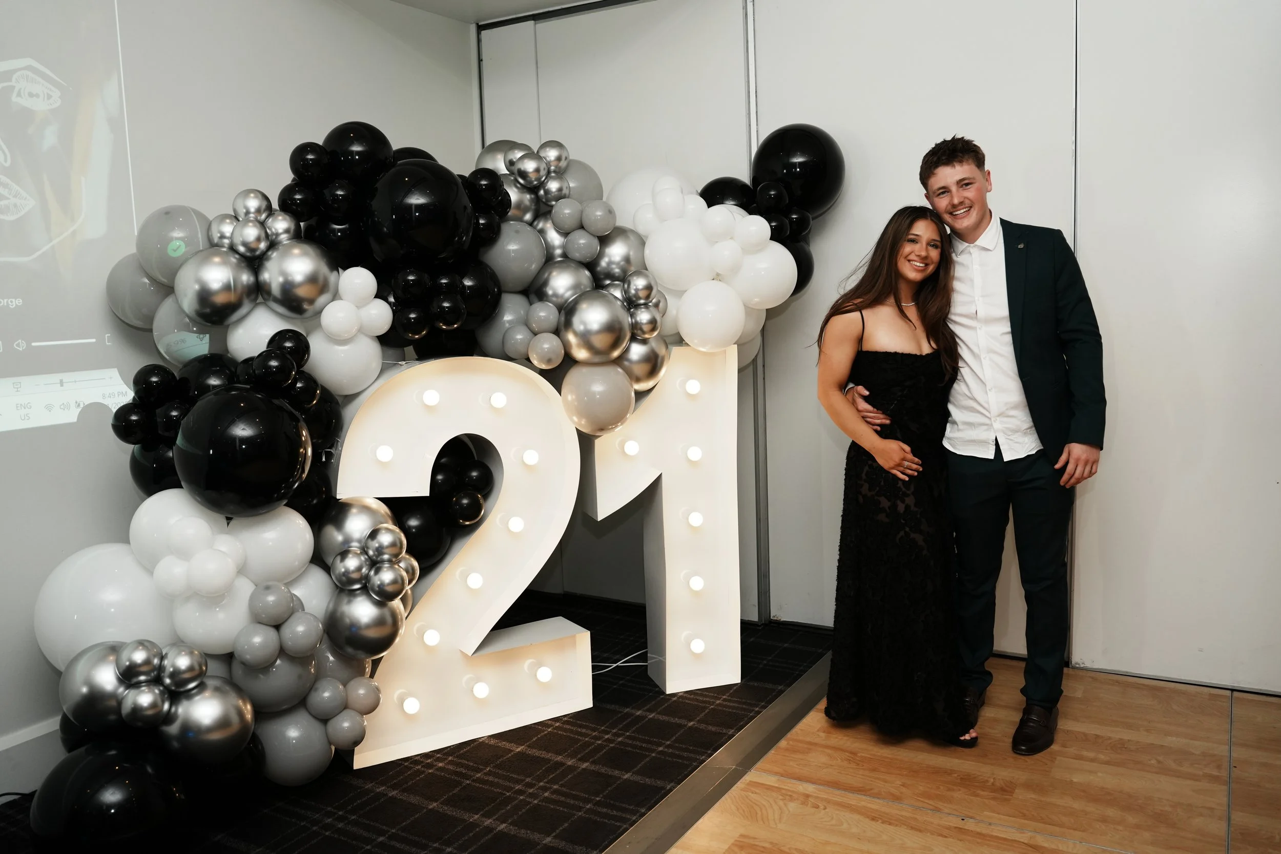 A couple dressed in formal attire standing next to large illuminated number 20 with a black, silver, gray, and white balloon arrangement celebrating a 20th anniversary or birthday.