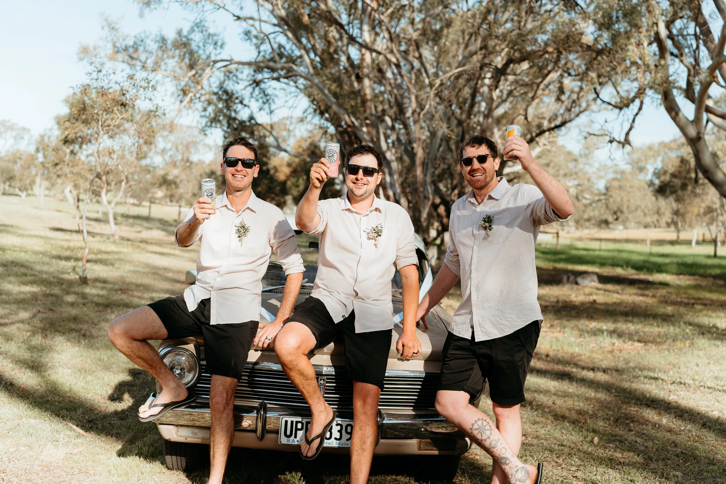 Three men in casual attire with sunglasses, sitting and standing on the front of a vintage car in a park-like setting, holding cans and smiling.