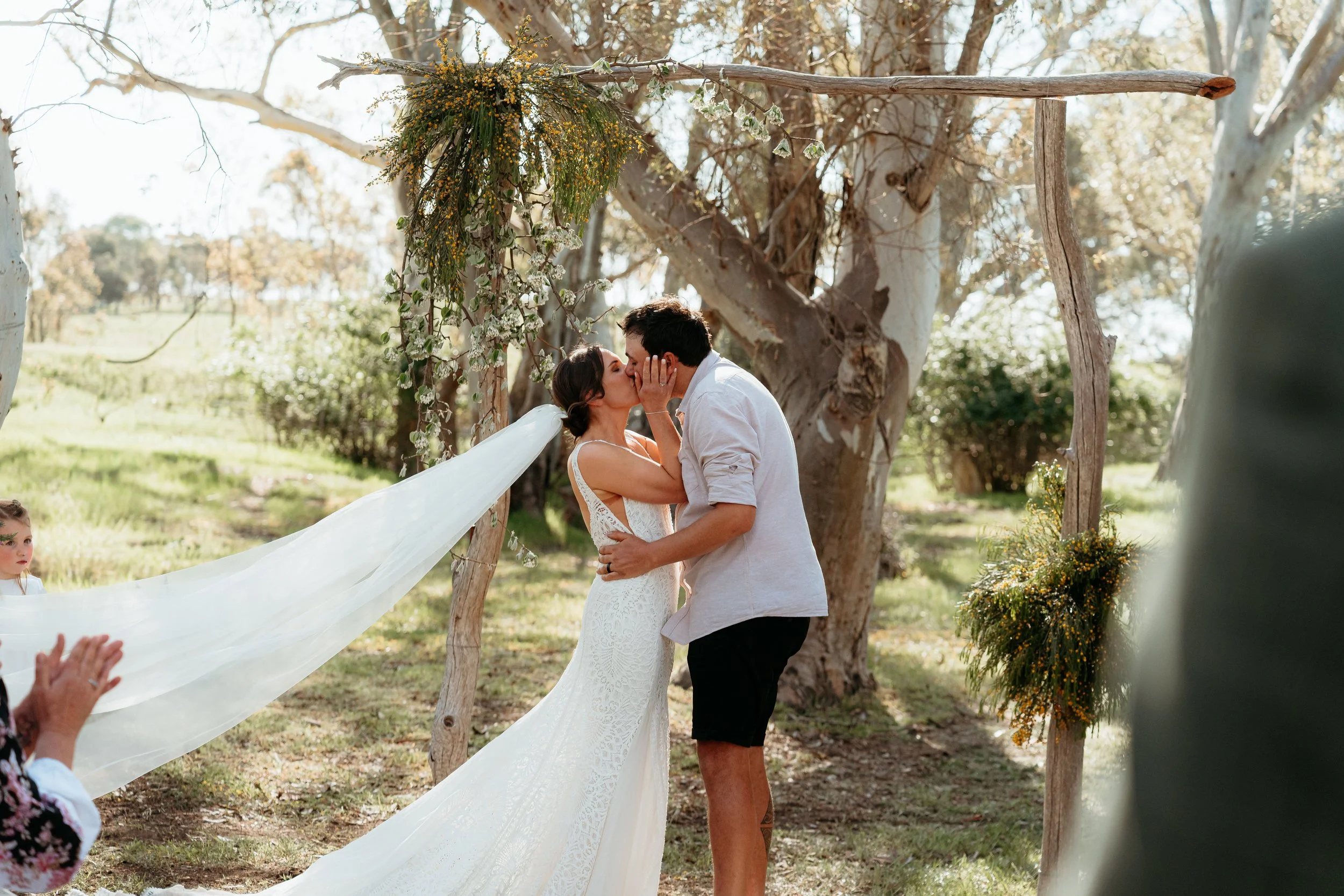 A bride and groom kiss during an outdoor wedding ceremony under a wooden arch decorated with flowers and greenery, with trees and sunlight in the background.