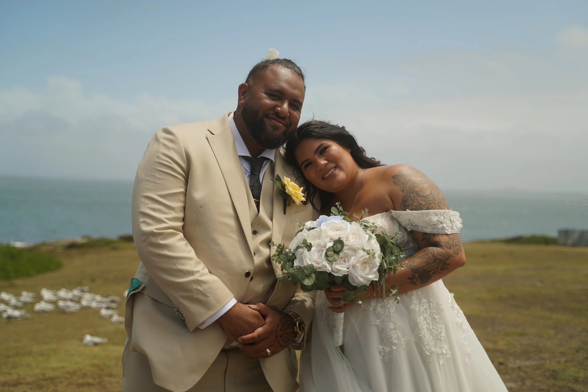 A newlywed couple standing outdoors on a grassy area with the ocean in the background, smiling and holding a bouquet of white roses.