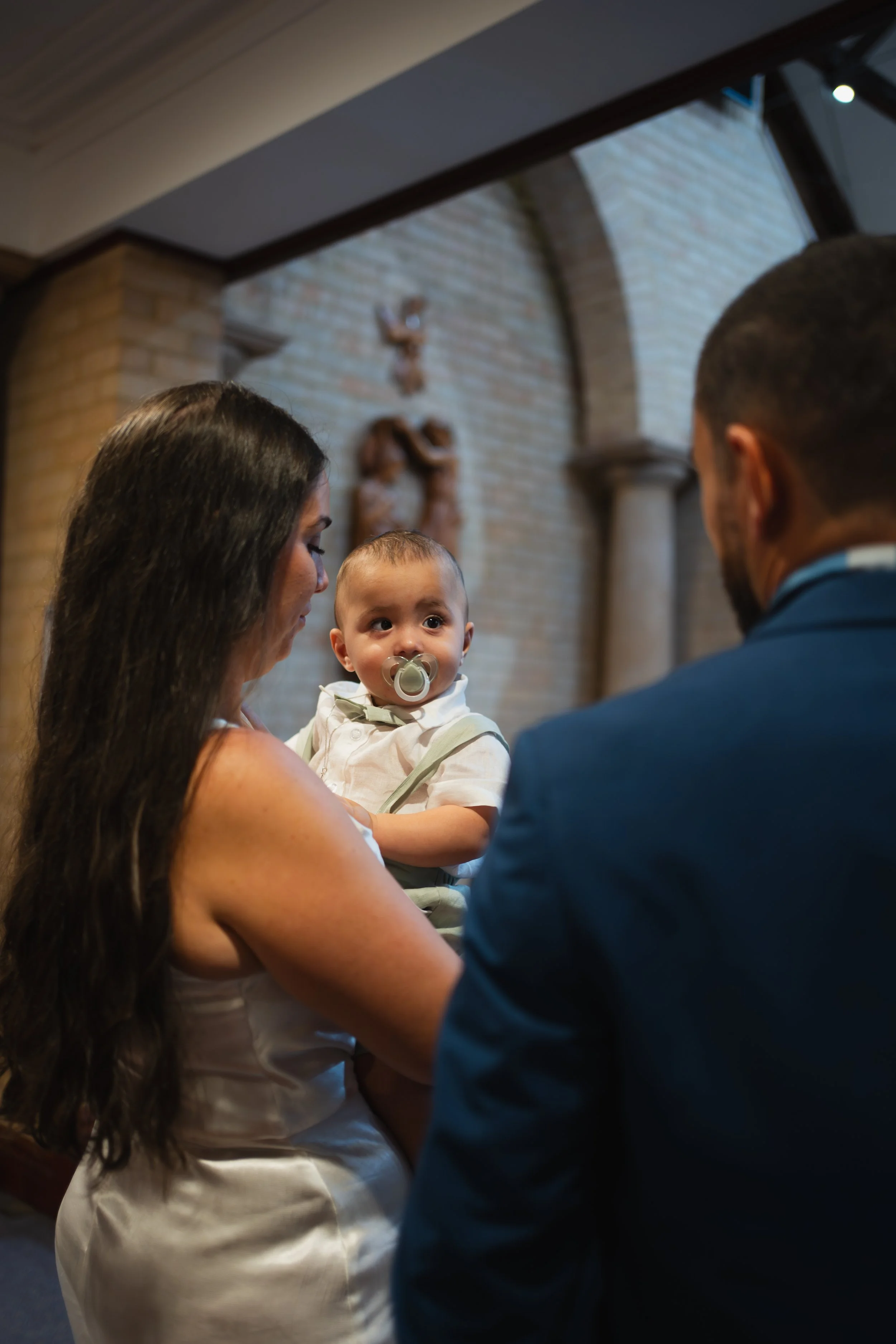 A woman holding a young boy with a pacifier in his mouth, facing a man in a blue suit, inside a church with religious sculptures on the brick wall behind them.