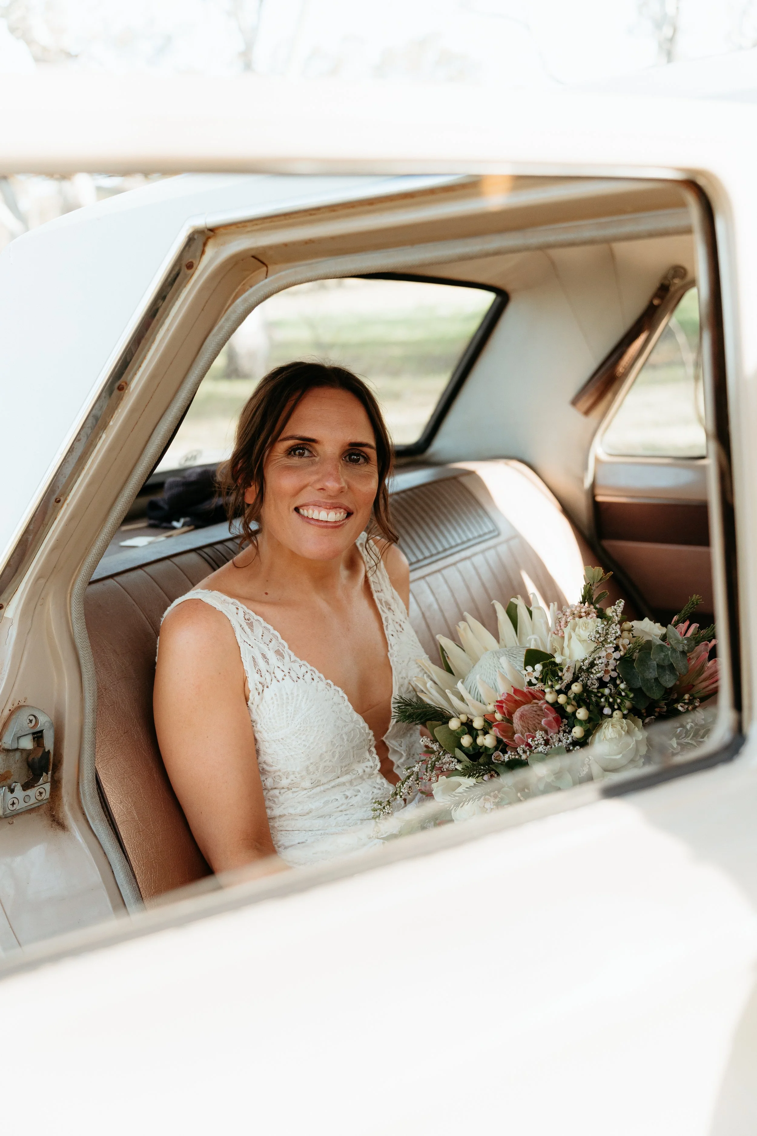 A woman dressed in a white lace wedding gown sitting inside a vintage car, holding a bouquet of flowers, smiling at the camera.