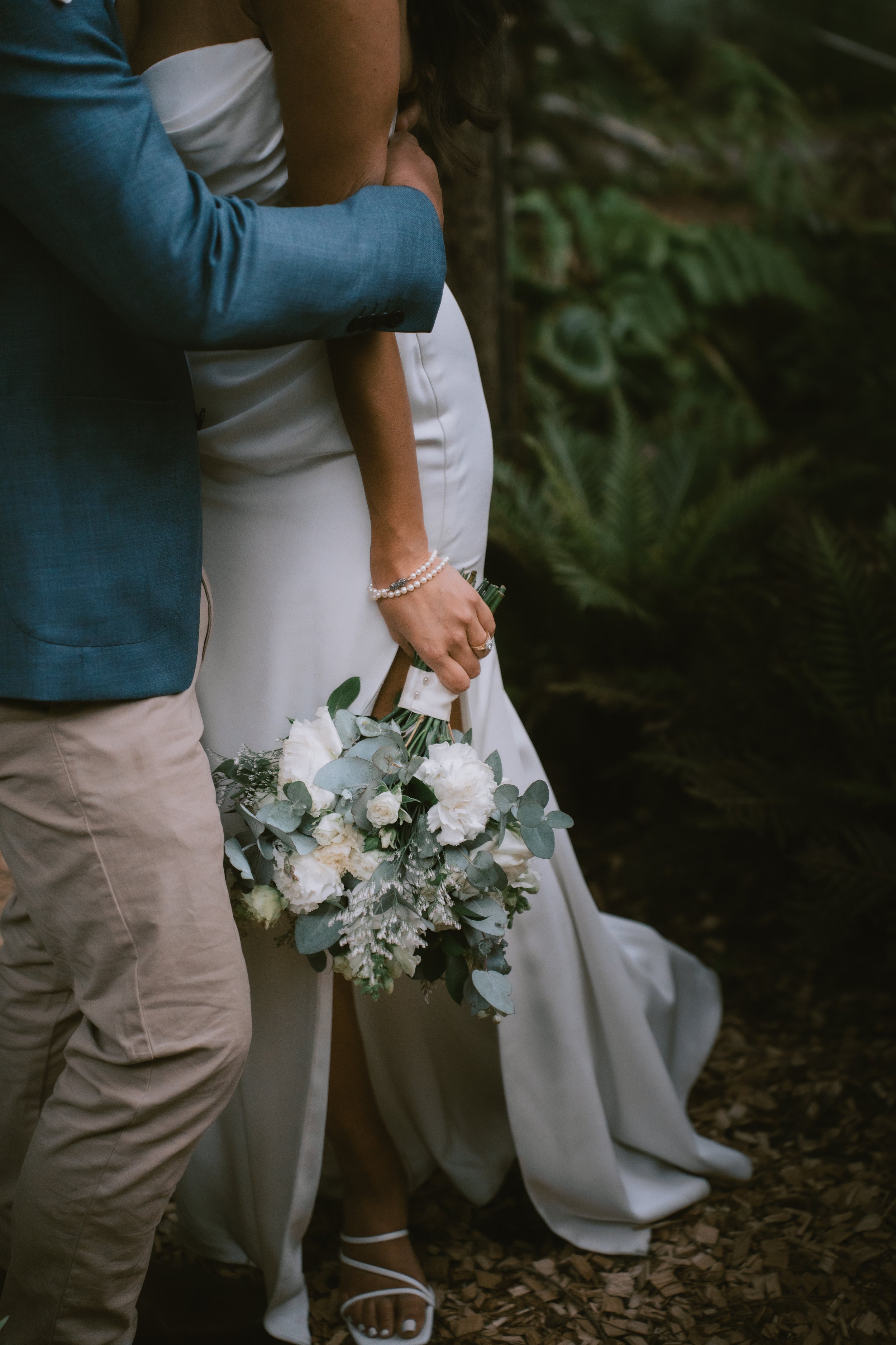 Bride holding a bouquet of white flowers and greenery while standing on a wooded trail, wearing white heels and a long white dress, with a man in a blue jacket and beige pants beside her.