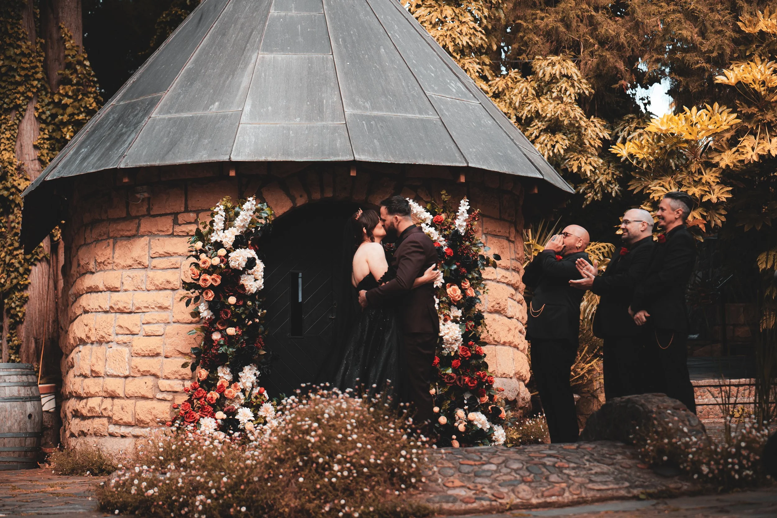 A couple kisses during their wedding ceremony outside a small stone chapel decorated with flowers. Two men in formal suits watch and applaud.