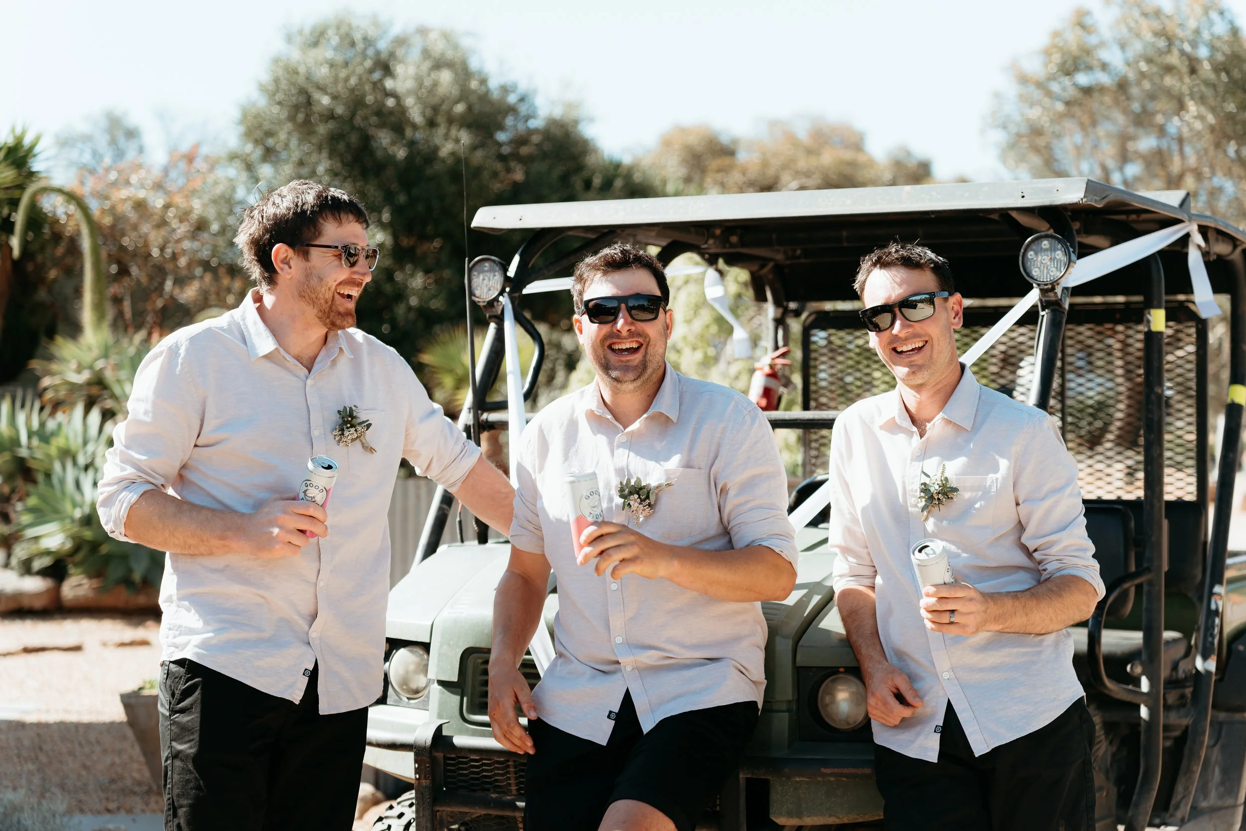 Three men wearing white shirts and sunglasses, smiling and holding drinks, standing next to a golf cart outdoors on a sunny day.