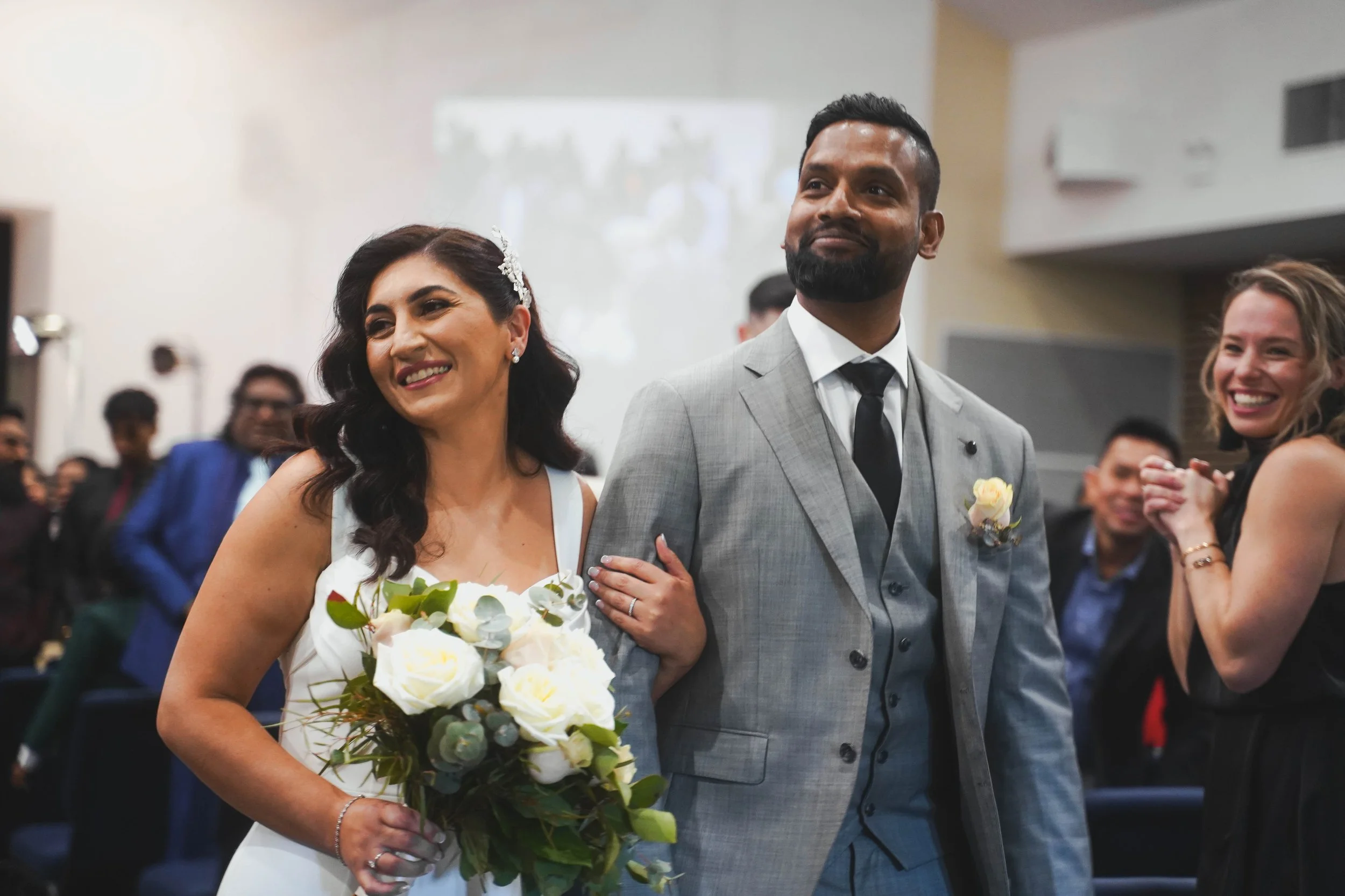 Bride and groom walking down the aisle holding hands in a wedding ceremony, with guests clapping and smiling in the background.