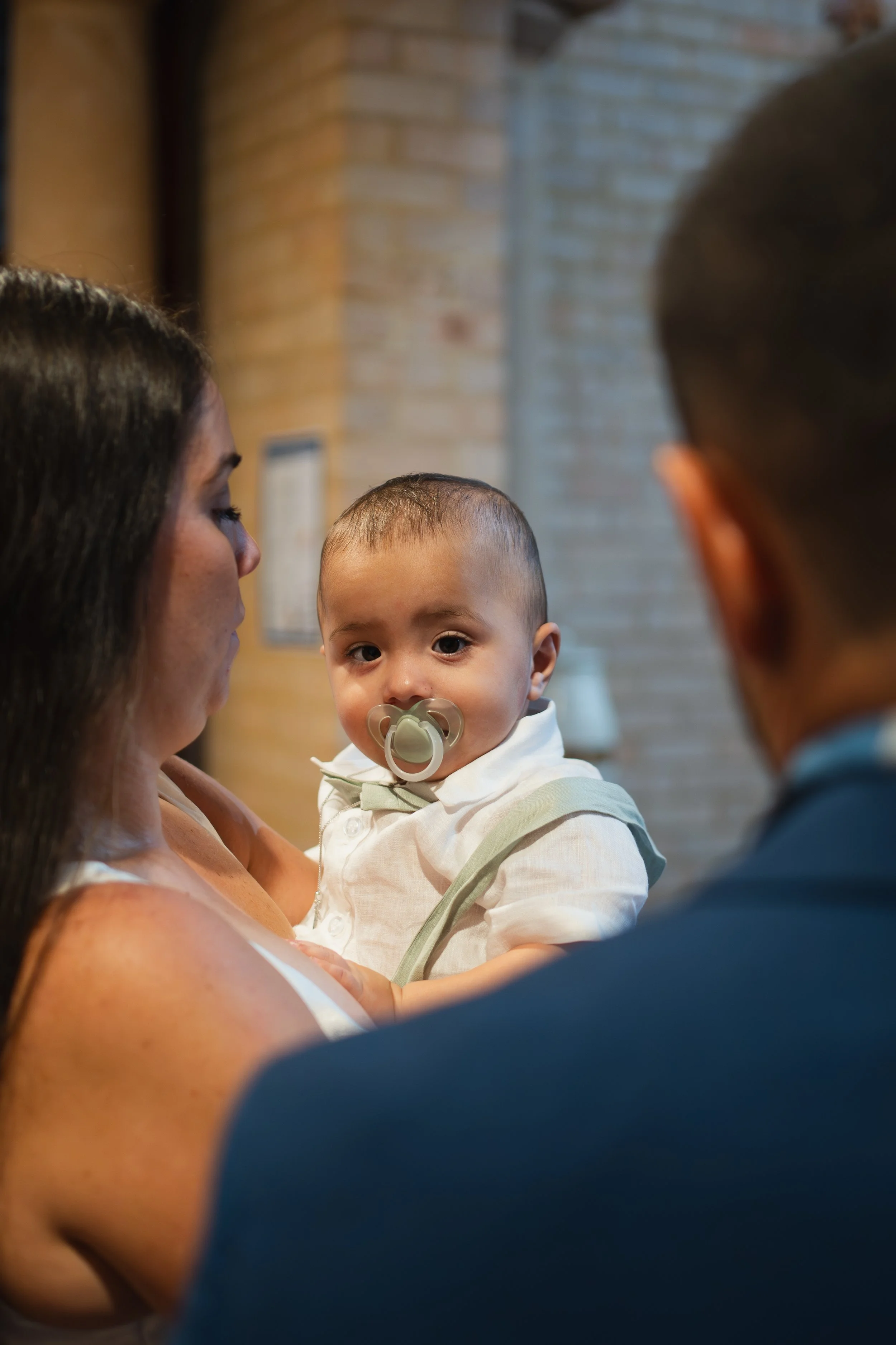 A woman holding a young boy with a pacifier in his mouth, engaged in a conversation with a man whose face is not visible. Background includes a brick wall.