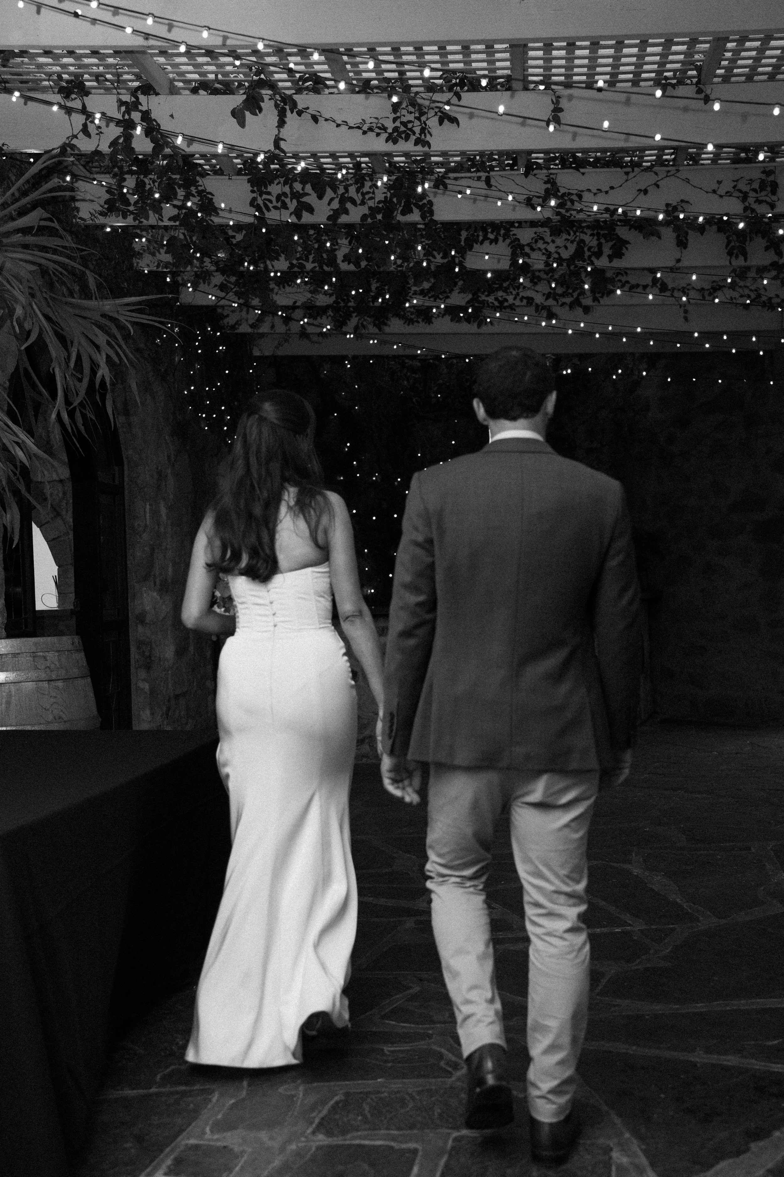 A bride and groom holding hands walking under string lights at night.