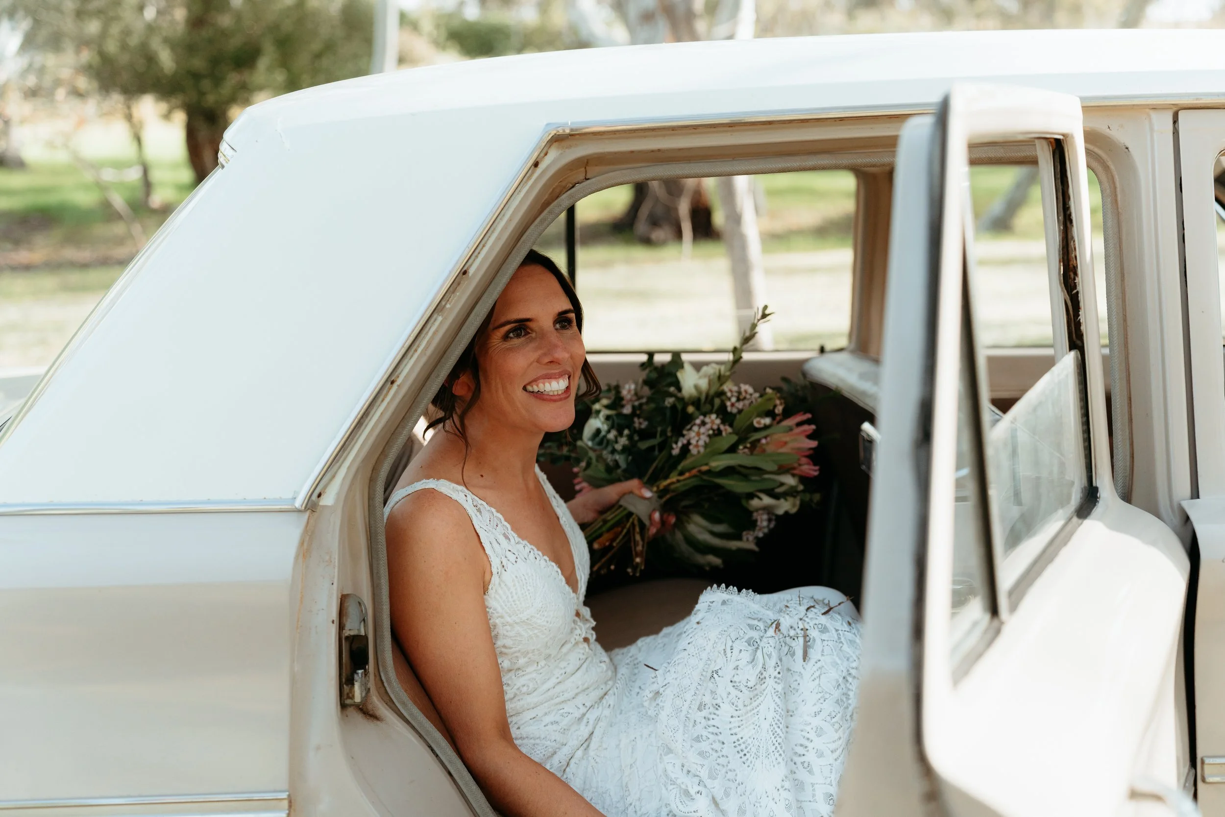 A smiling woman in a white lace dress sitting inside a vintage cream-colored car with an open door, holding a bouquet of flowers, outdoors on a sunny day.