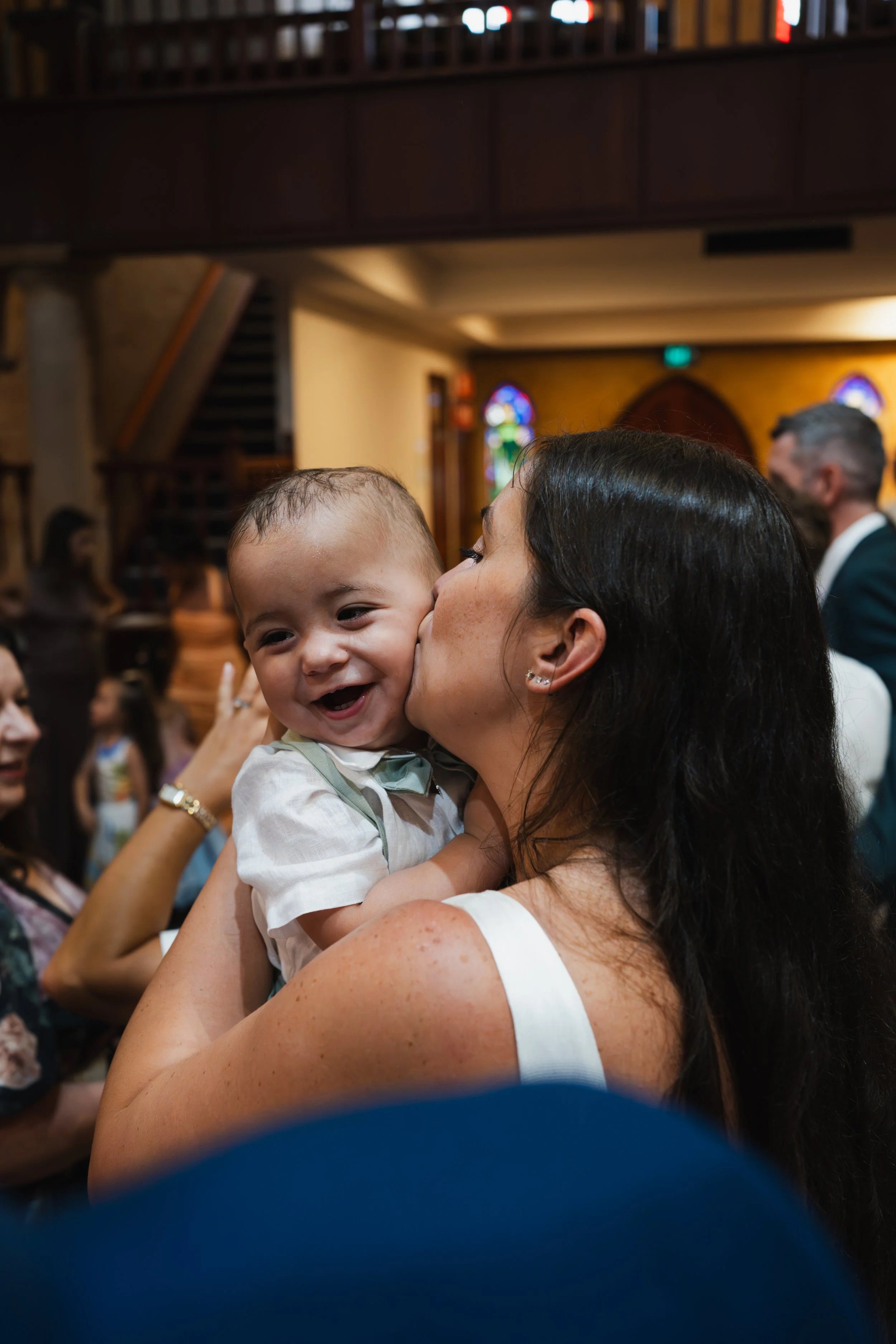 A woman is holding and kissing a smiling baby boy at an indoor event or celebration.