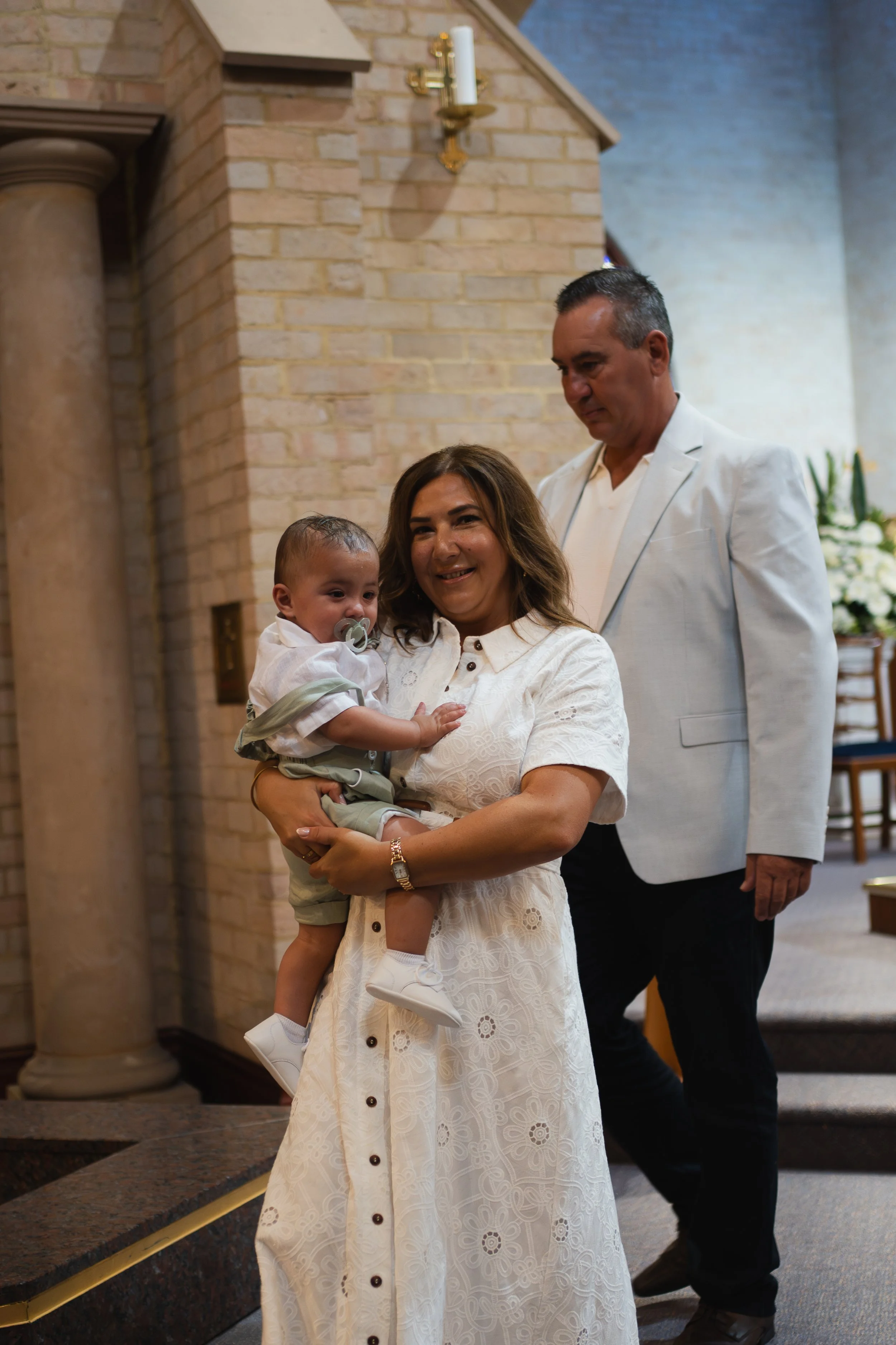 A woman holding a baby, with a man standing behind them in an indoor setting near a brick fireplace.
