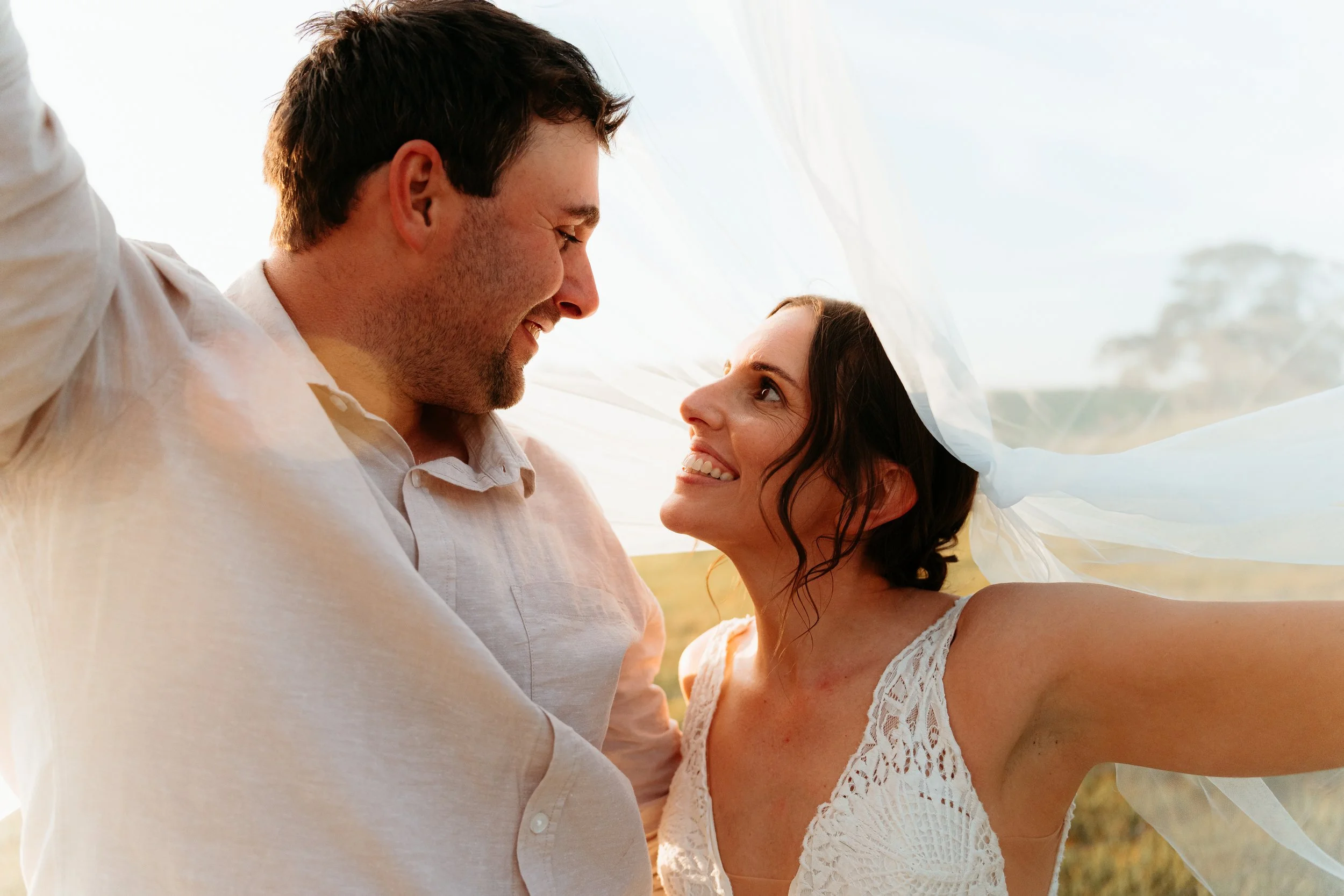 A couple smiling happily under a sheer white cloth outdoors during sunset.