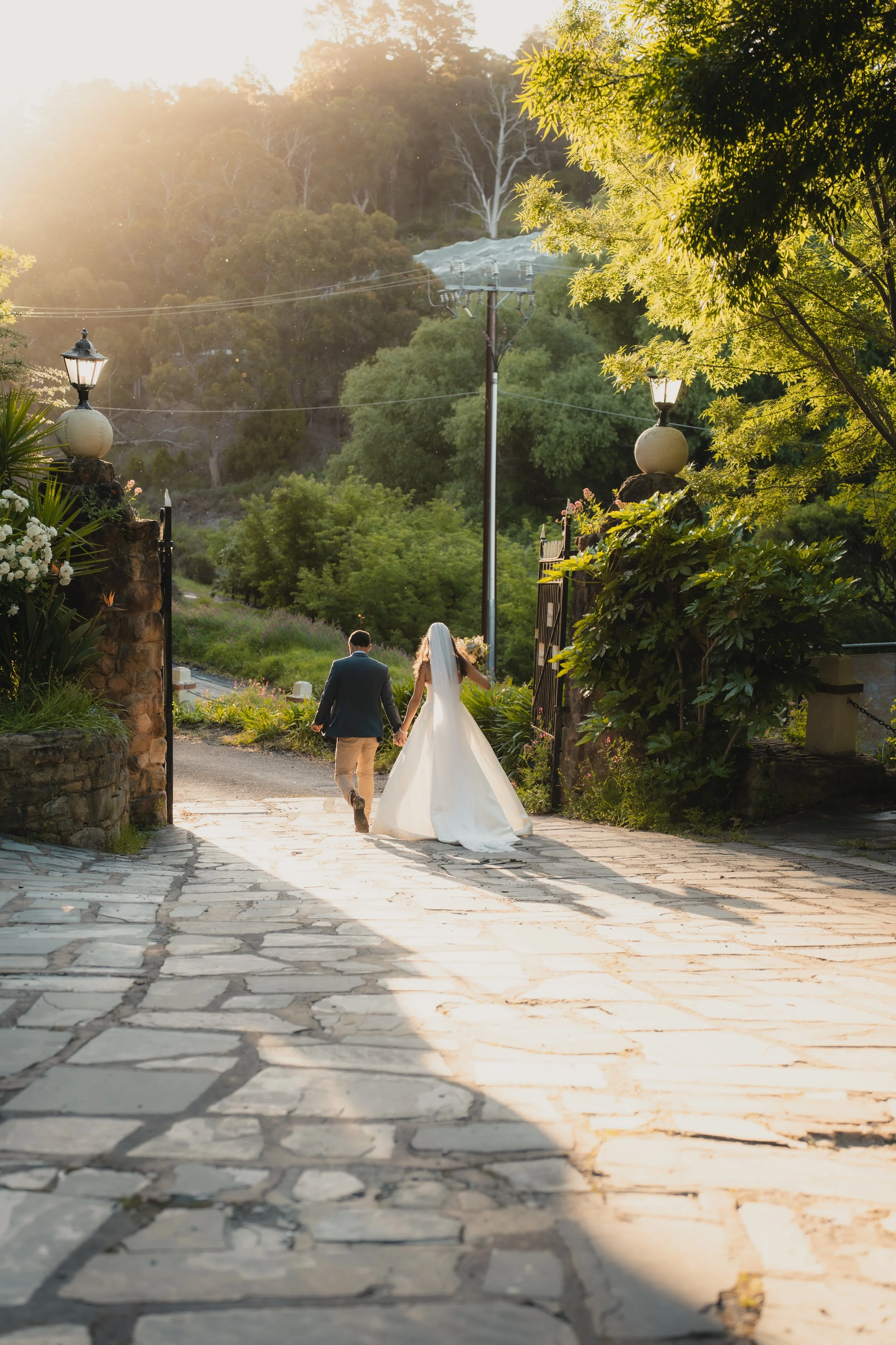 A bride and groom walking hand-in-hand through an arched stone gate during sunset, with lush green trees and mountains in the background.