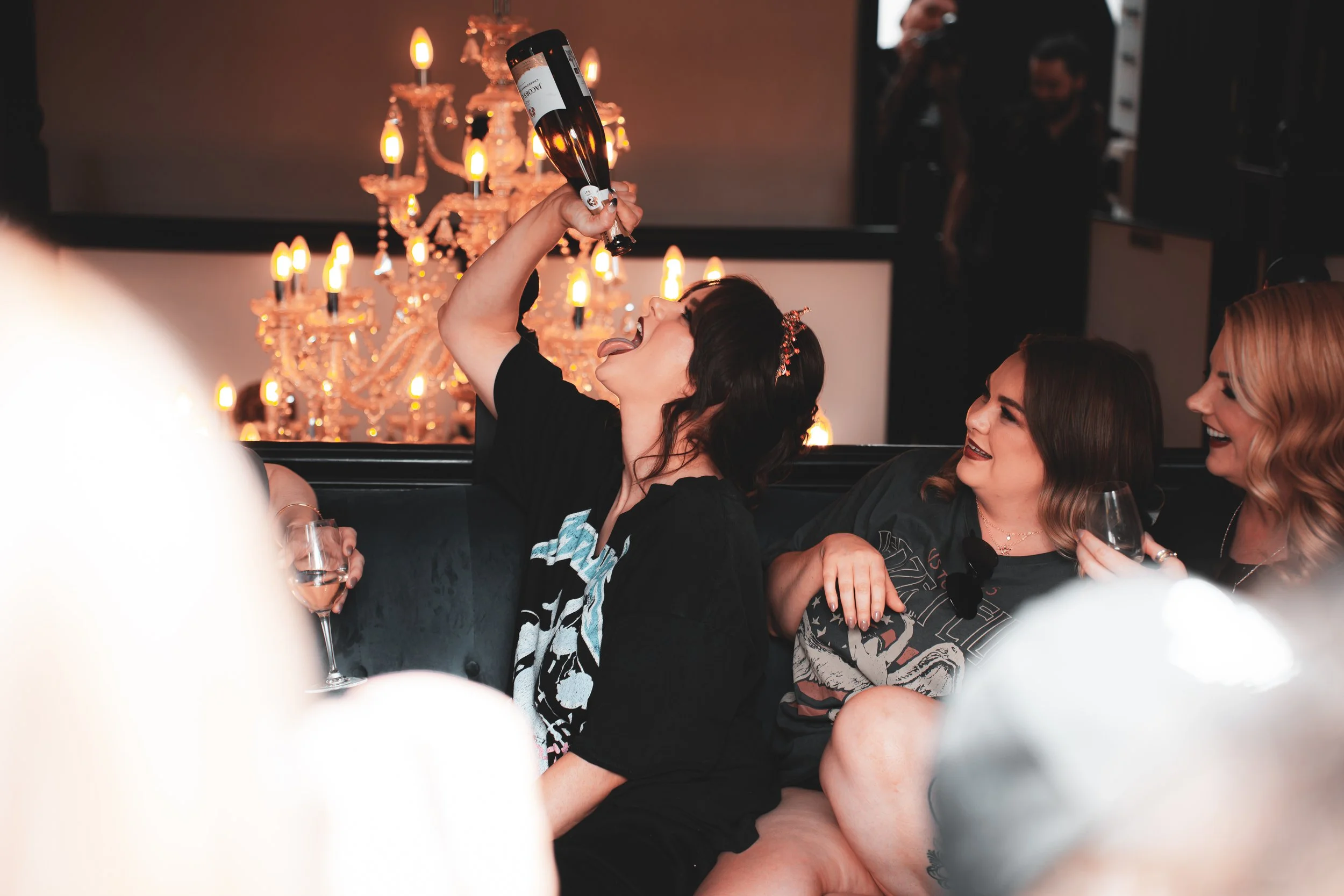 Woman wearing black shirt holding a wine bottle upside down over her tongue at a celebration, surrounded by friends smiling and holding glasses, elegant chandelier in the background.