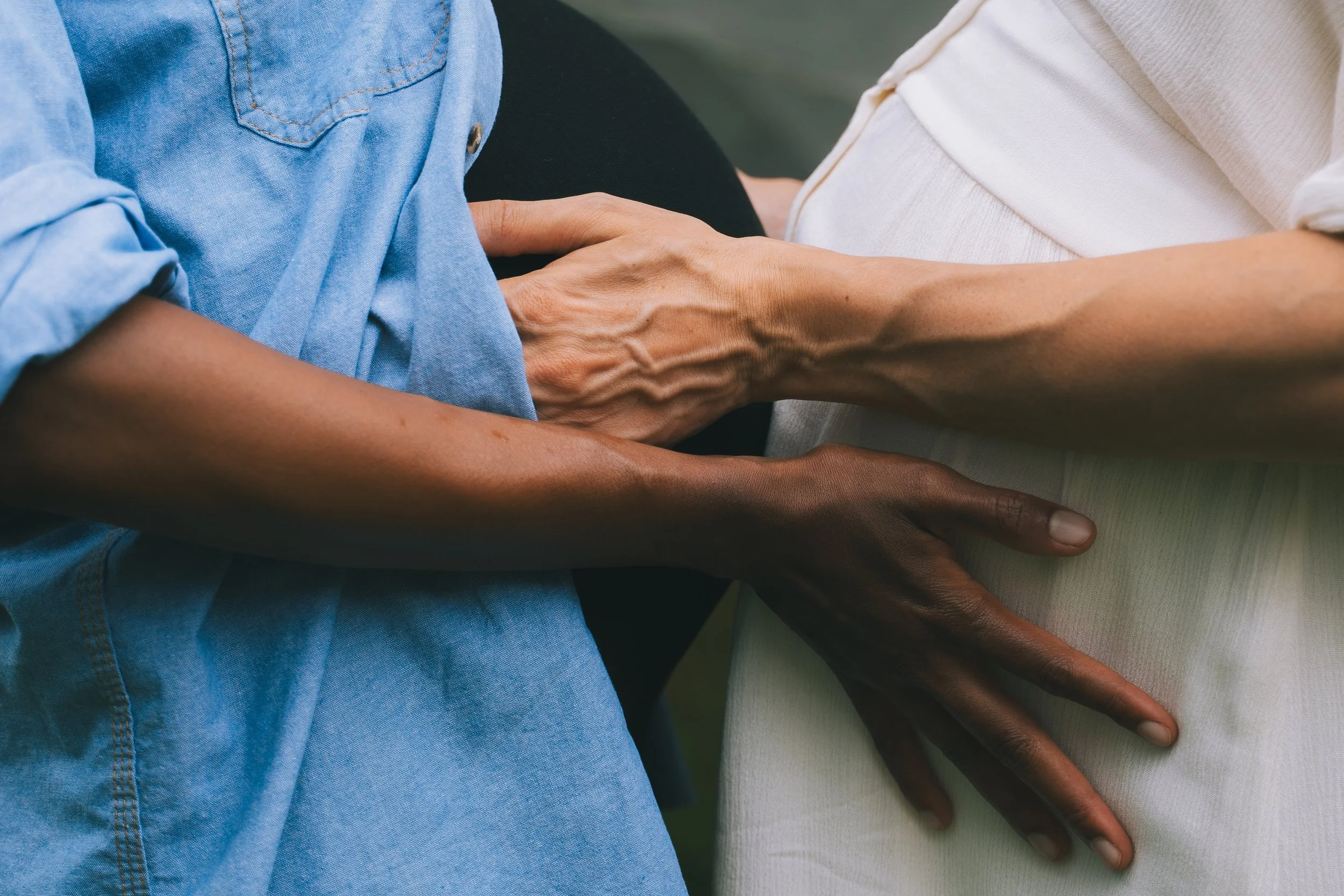 Close-up of three hands, one light-skinned, one dark-skinned, and one with veins visible, gently touching or holding each other in a gesture of support or connection.