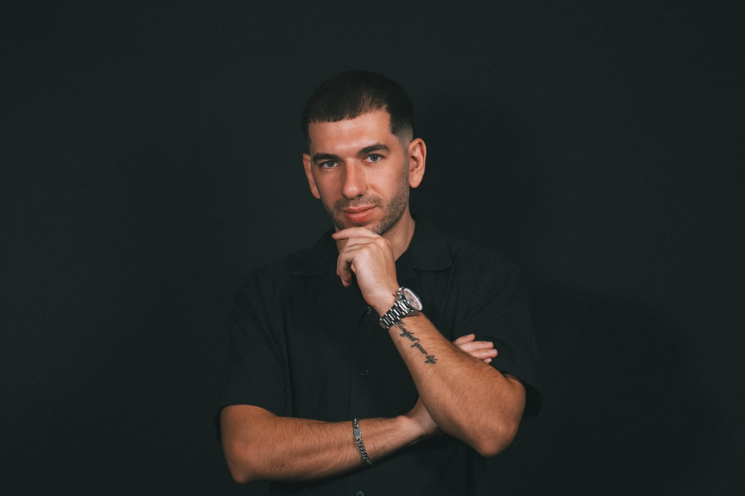 A young man with short dark hair and light facial hair posing with his chin resting on his hand, wearing a black shirt, a silver watch, and a bracelet, against a dark background.