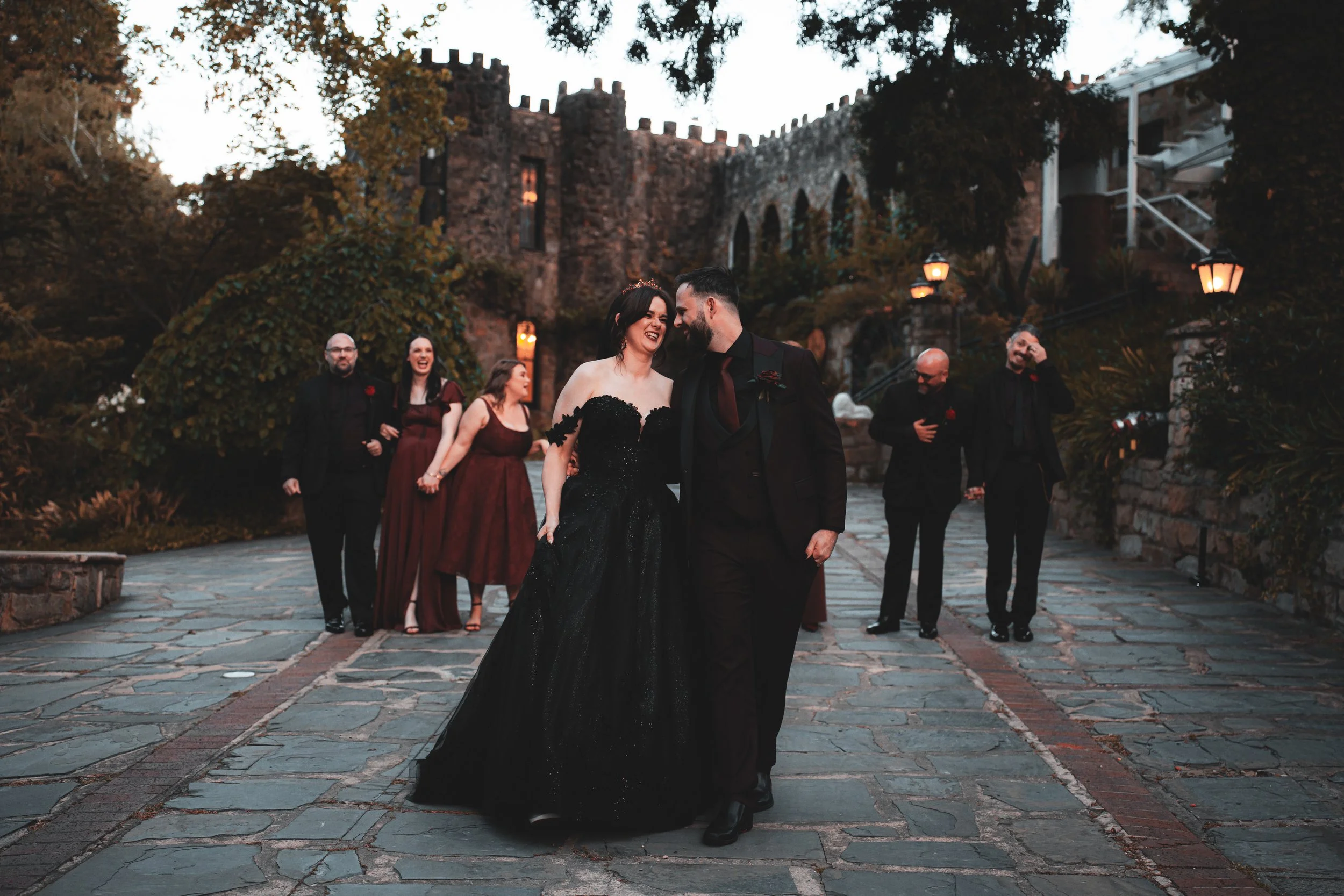 A joyful wedding couple walks hand in hand on a stone pathway, surrounded by friends and family, outside a castle-like stone building at dusk.