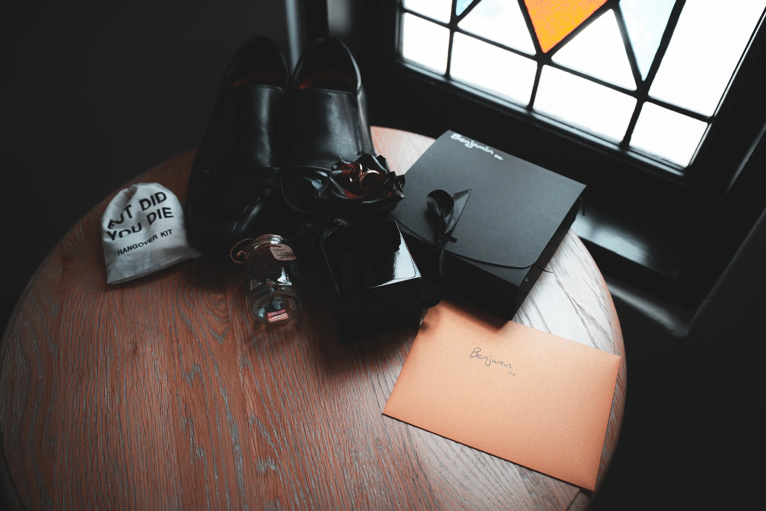 A round wooden table with a pair of black shoes, a white hangover kit bag that says 'What did you die', a black box, an orange envelope with the name 'Benjamin', a black notebook, a black box, a pair of glasses, and a small perfume bottle. The table 