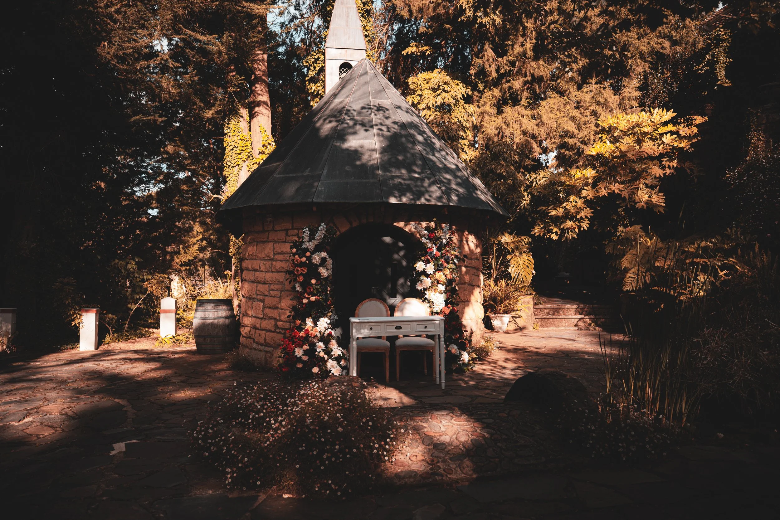 Small stone building with a conical metal roof, decorated with colorful flowers around the entrance, surrounded by trees and plants, with stone steps leading up to it.