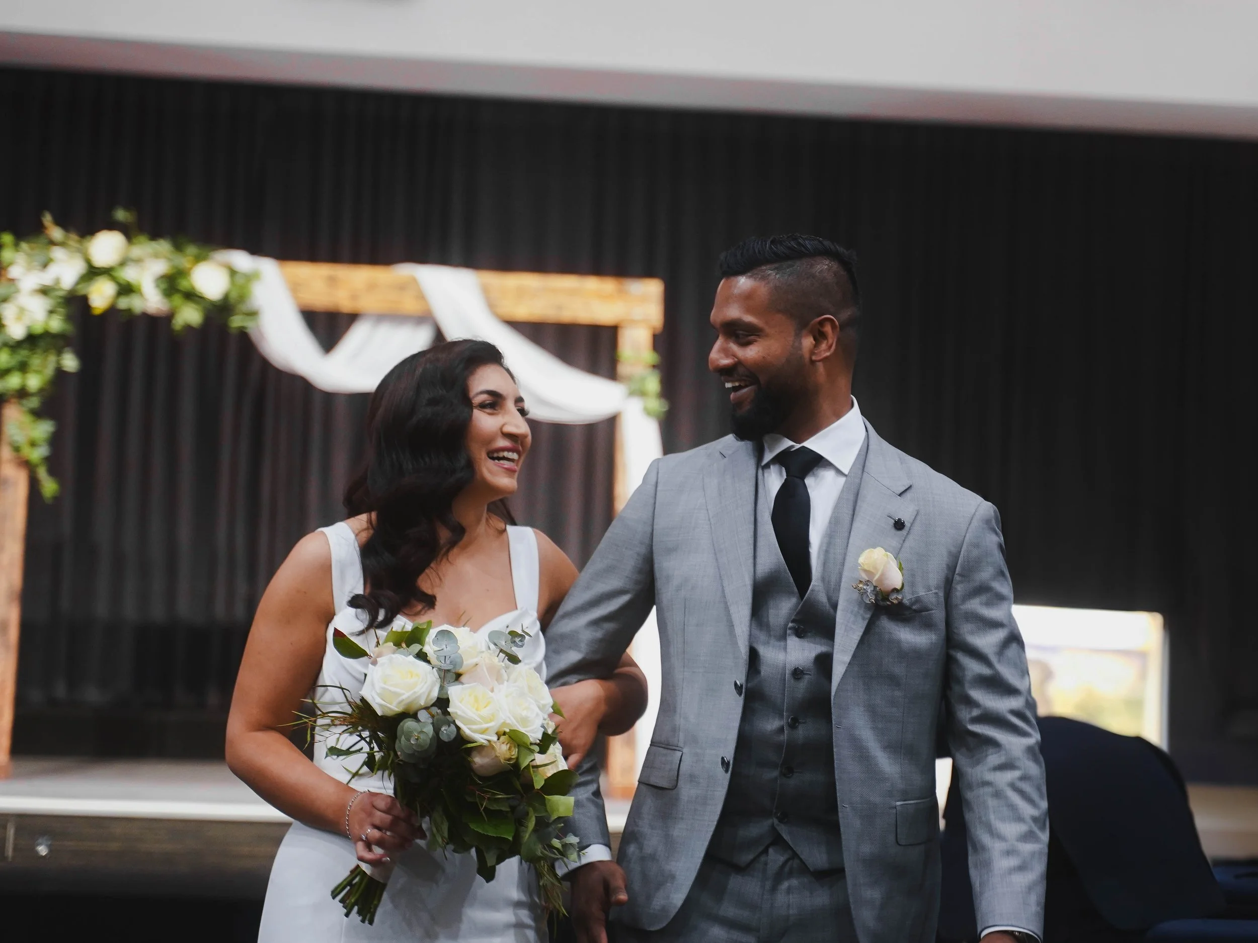 A happy bride and groom smiling at each other during their wedding ceremony, with a floral and fabric arch in the background.