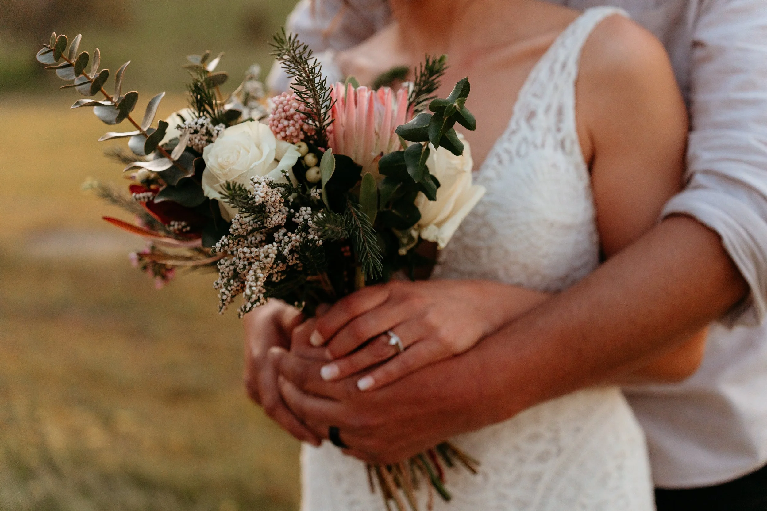 A bride in a white lace dress holding a bouquet of flowers, with a groom behind her embracing her. The bouquet includes white roses, pink protea, greenery, and small white flowers.