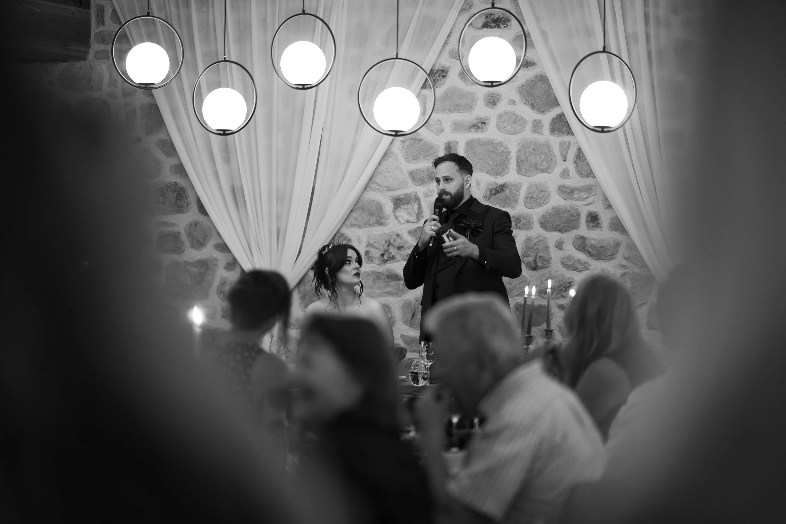 A man giving a speech at a wedding reception, with a woman sitting behind him, inside a decorated venue with stone walls and hanging lights.