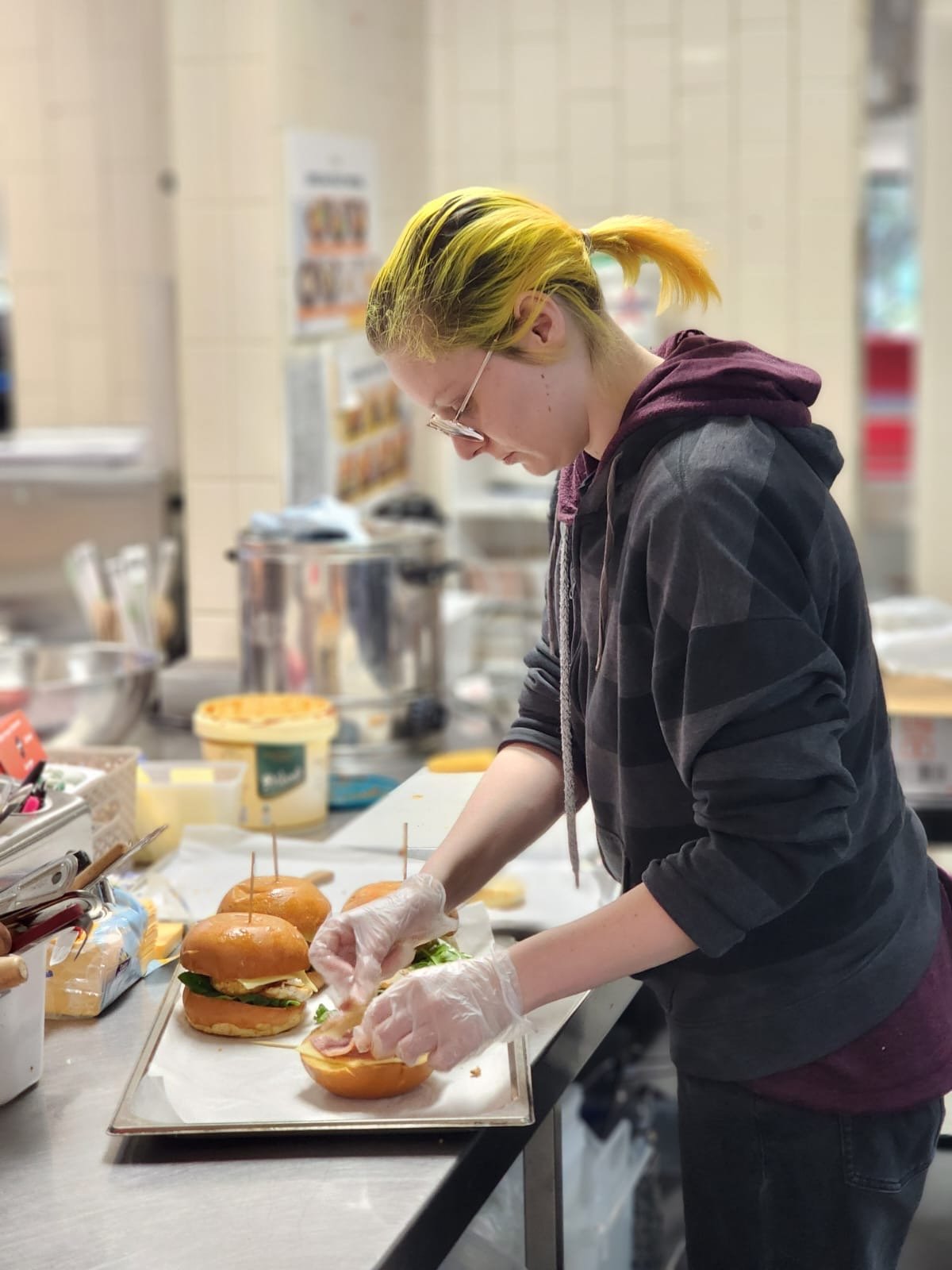 A person with yellow hair and glasses preparing sandwiches in a kitchen or cafeteria setting.
