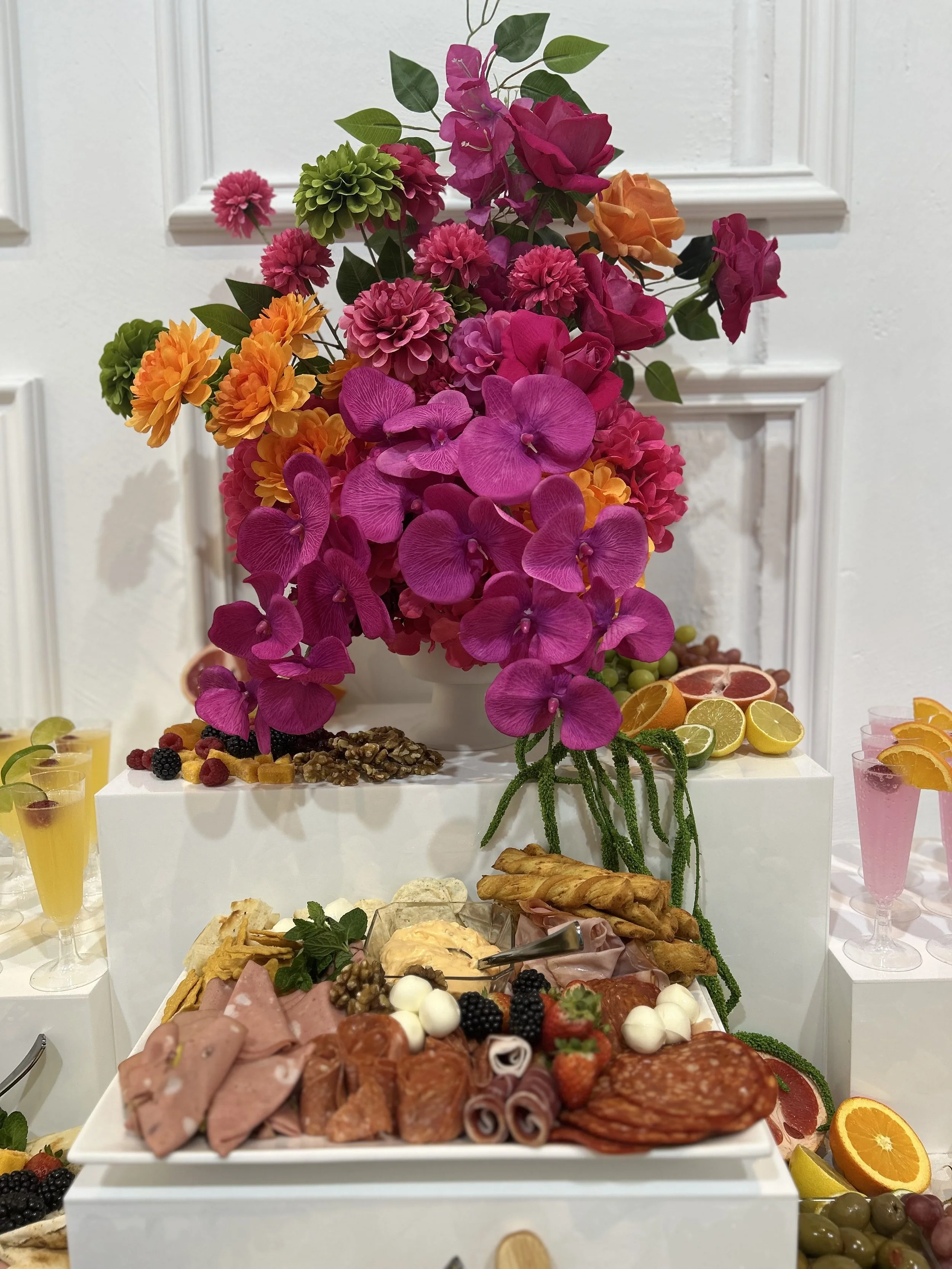 An elegant display of a large bouquet featuring pink, orange, and green flowers positioned on a white pedestal table. In front of the flowers, there is an assortment of sliced fruits, cured meats, cheeses, nuts, and garnishes, along with glasses of c