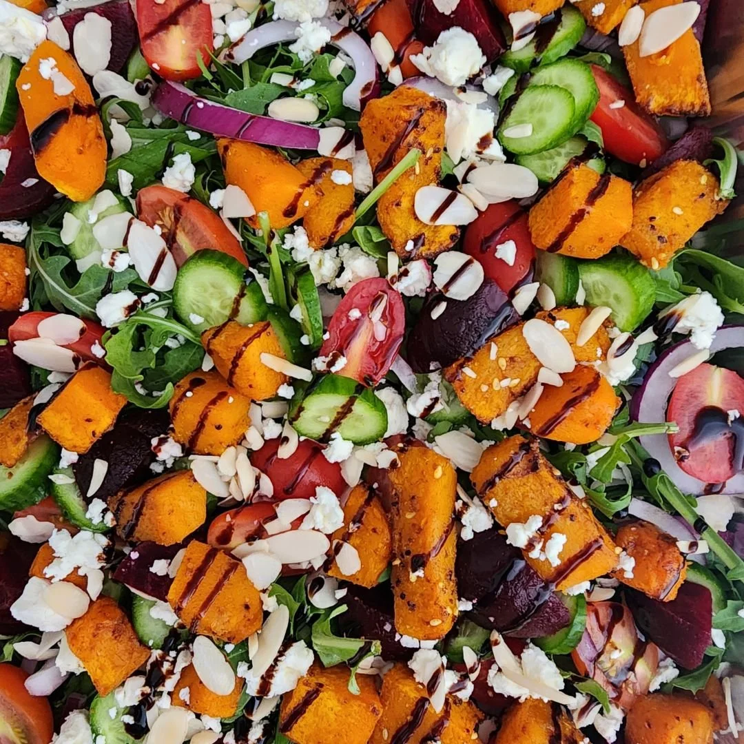 A colorful salad with roasted sweet potatoes, cherry tomatoes, cucumbers, red onions, greens, almond slices, feta cheese, and a drizzle of balsamic glaze.