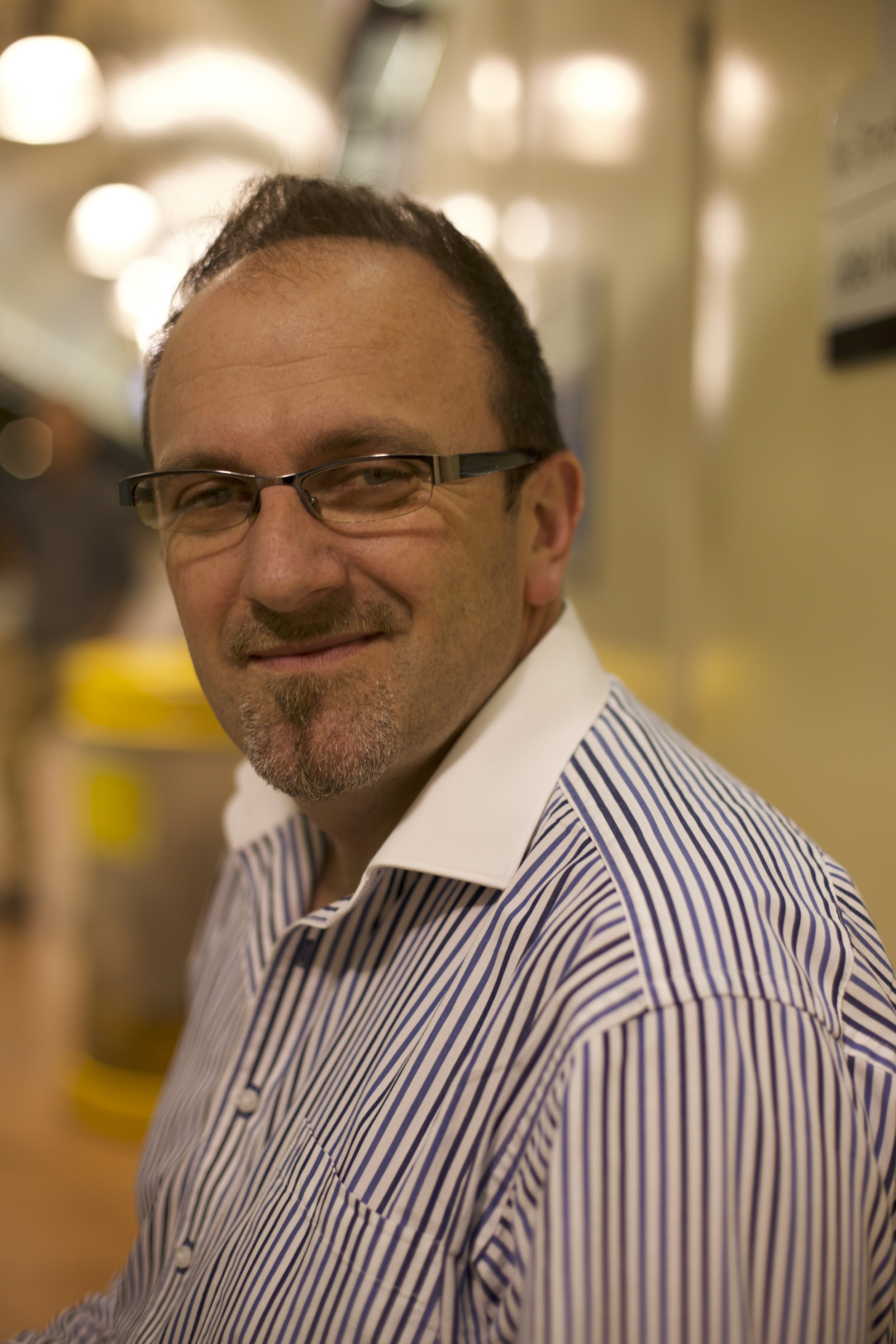A man wearing glasses and a striped shirt sitting indoors with warm lighting, smiling at the camera.