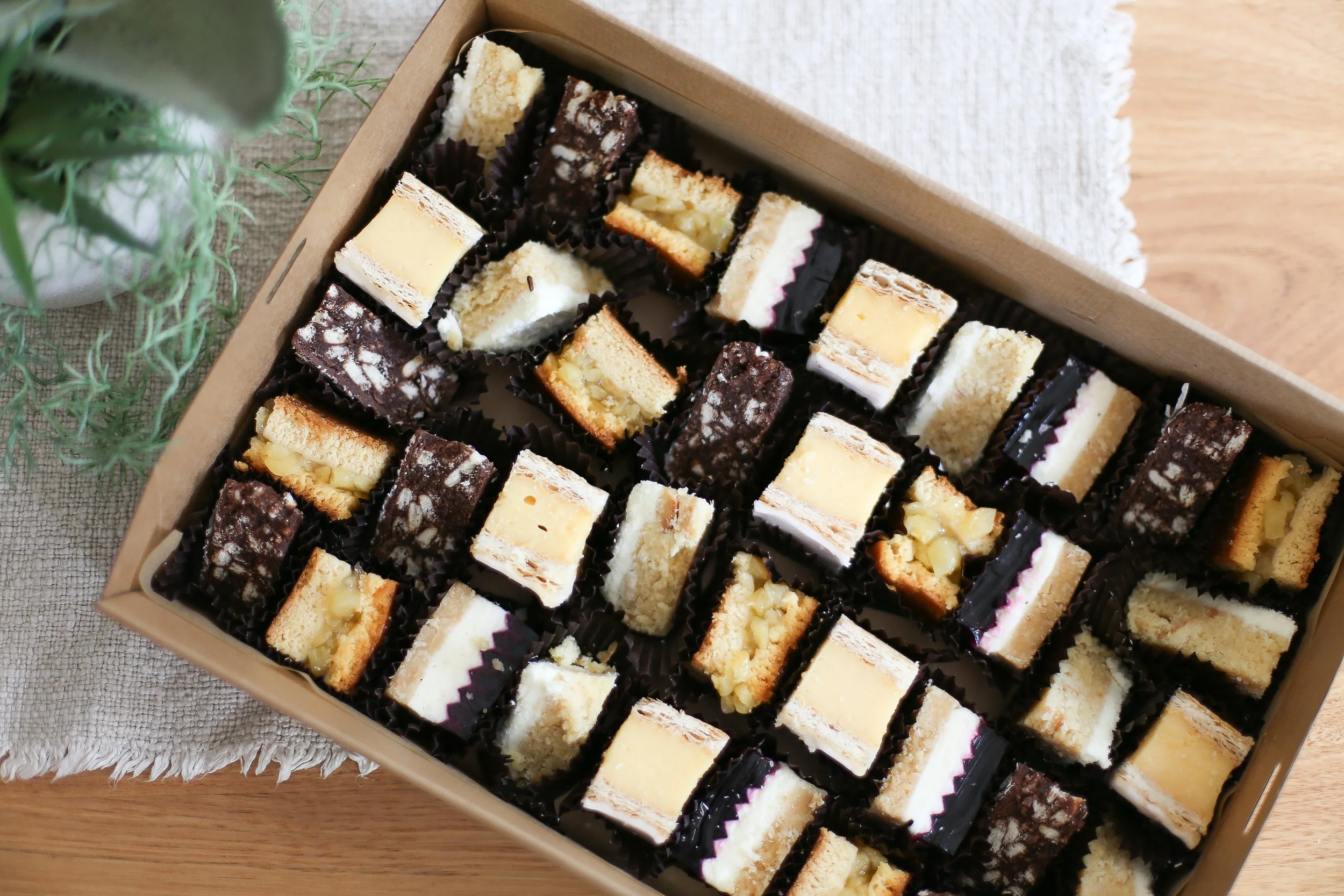 A box of assorted small dessert bars, including cheesecakes, layered cakes, and nut-topped bars, arranged in black paper cups on a wooden surface.