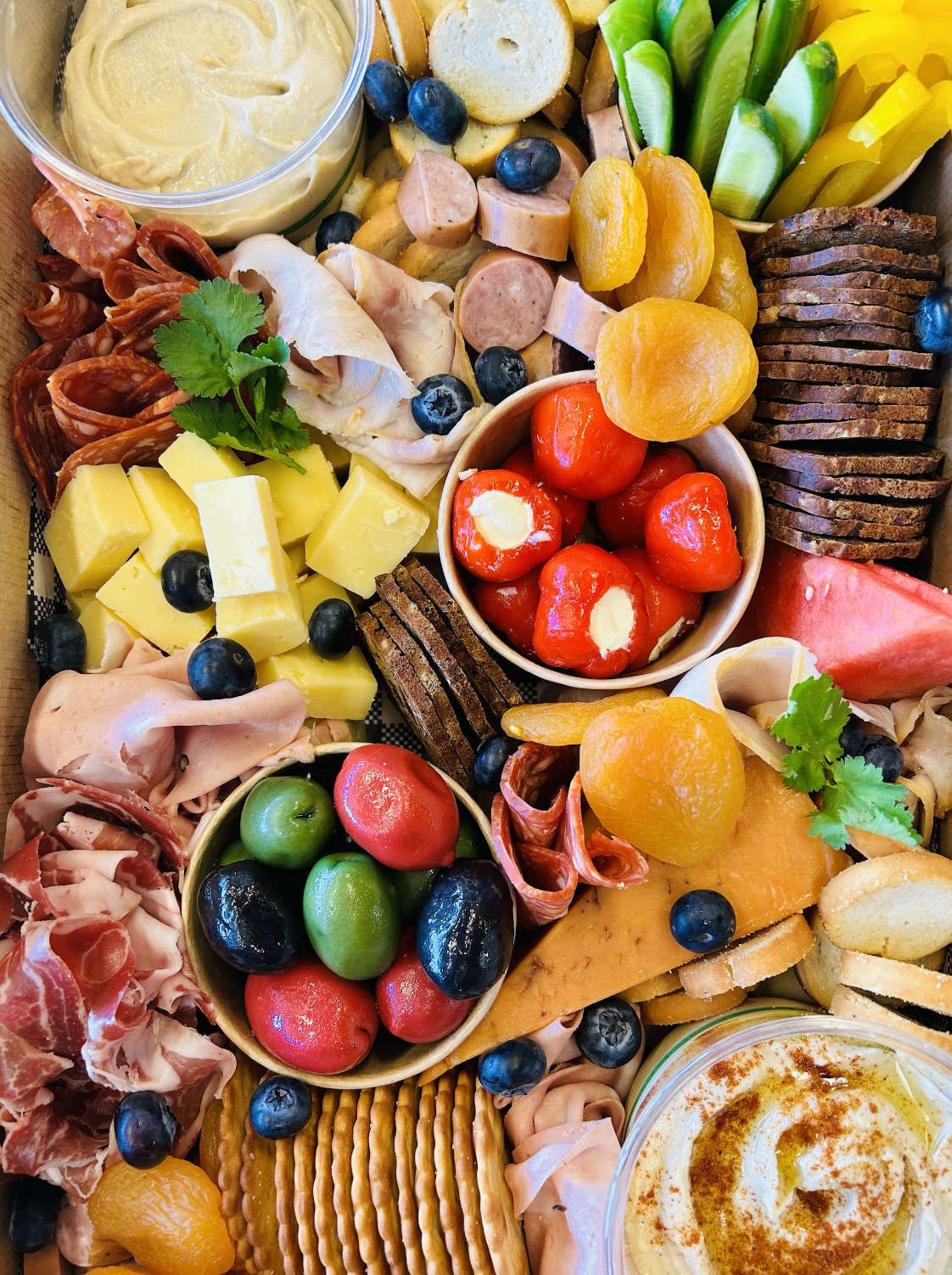 A variety of snacks including cheese cubes, grapes, blueberries, sliced meats, crackers, cherry peppers, and sauces.