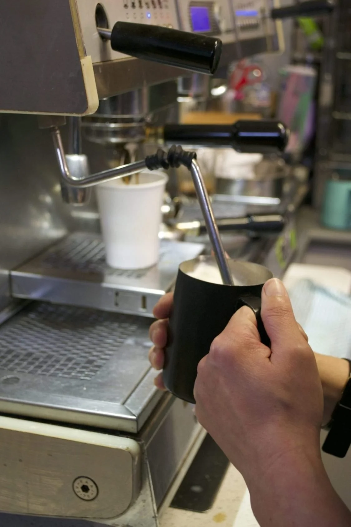 Person holding a black mug under a coffee machine's steam wand.