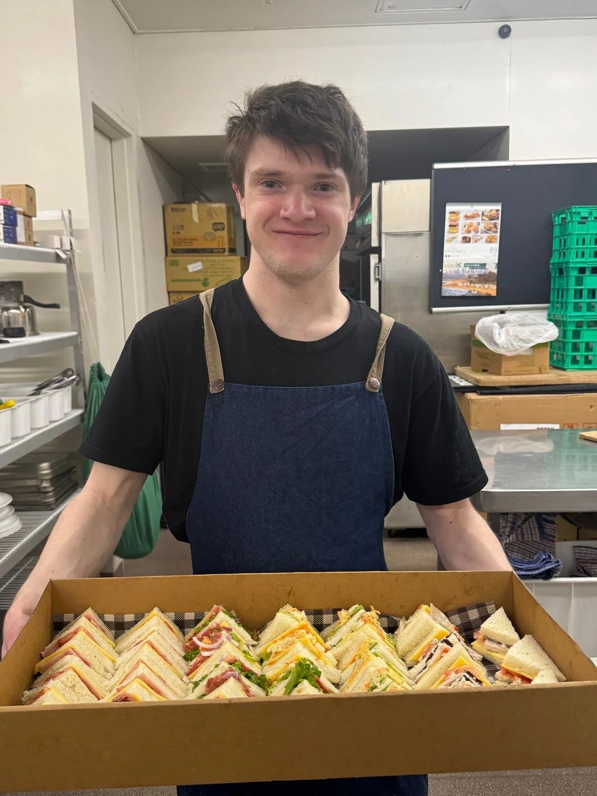 A young man in a black T-shirt and apron holding a tray of assorted sandwiches in a kitchen setting.
