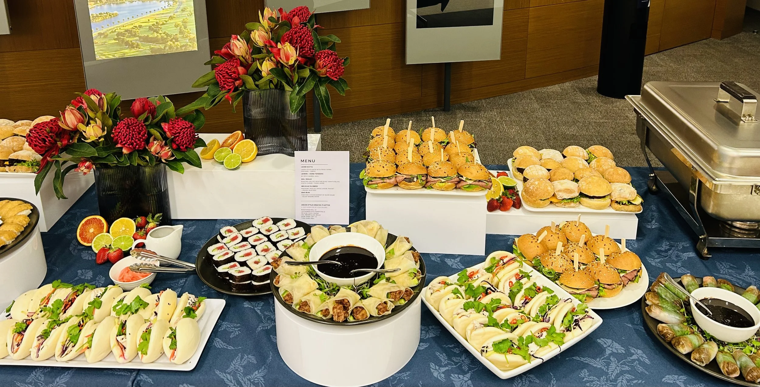 Assorted food items on a buffet table, including mini sandwiches, sushi rolls, fruit slices, and salads, with floral centerpieces featuring red and pink flowers.