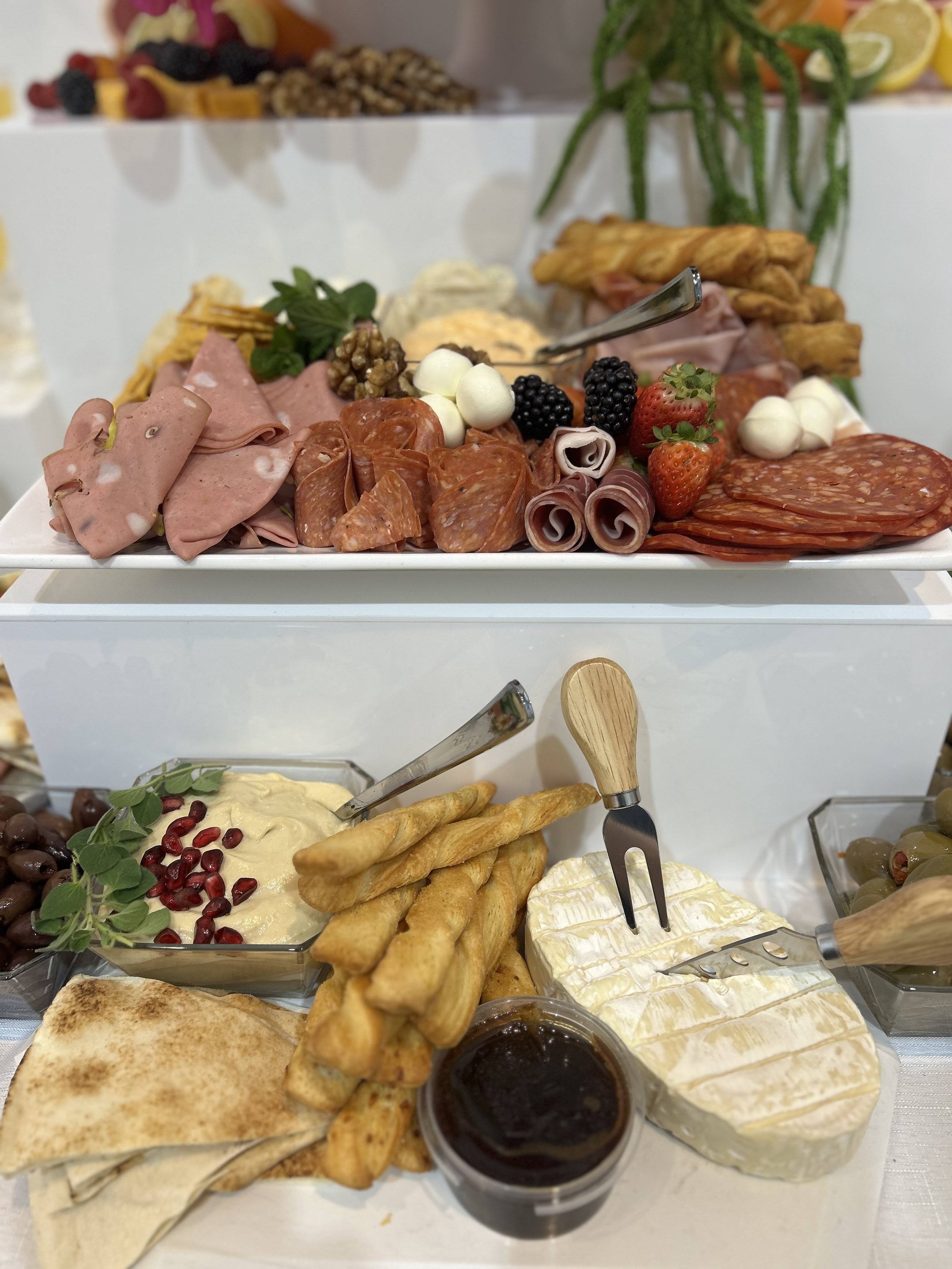 Cheese platter with brie, breadsticks, olives, grape jam, and other Mediterranean appetizers, with a second tier of assorted meats, fruits, cheeses, and crackers.