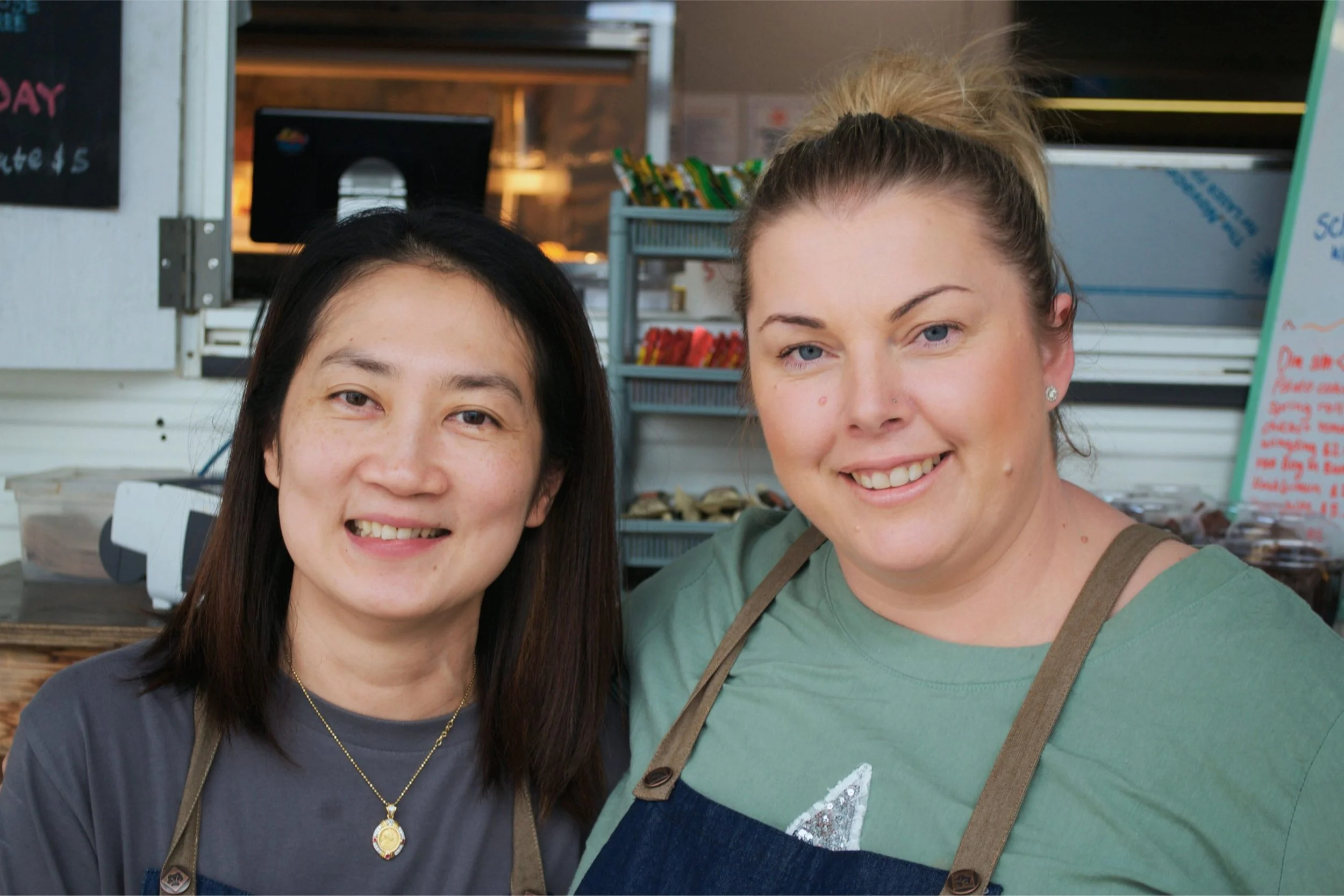 Two smiling women standing behind a food stand, one with black hair and the other with blonde hair tied up, in front of a menu board and snack shelves.