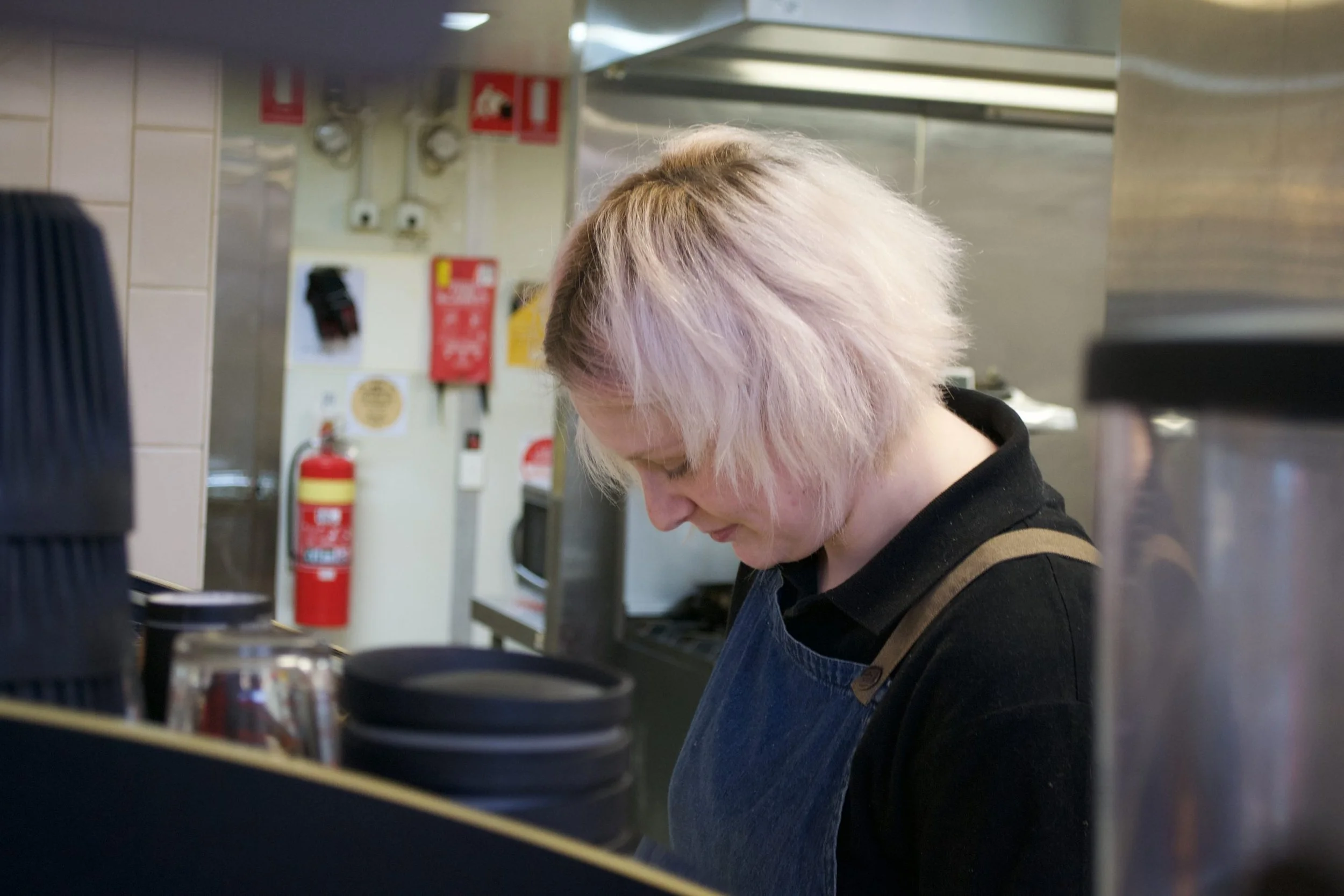 A woman with light, blond hair working in a kitchen, standing behind a counter with stacked black cups and glassware, with safety equipment and kitchen appliances in the background.