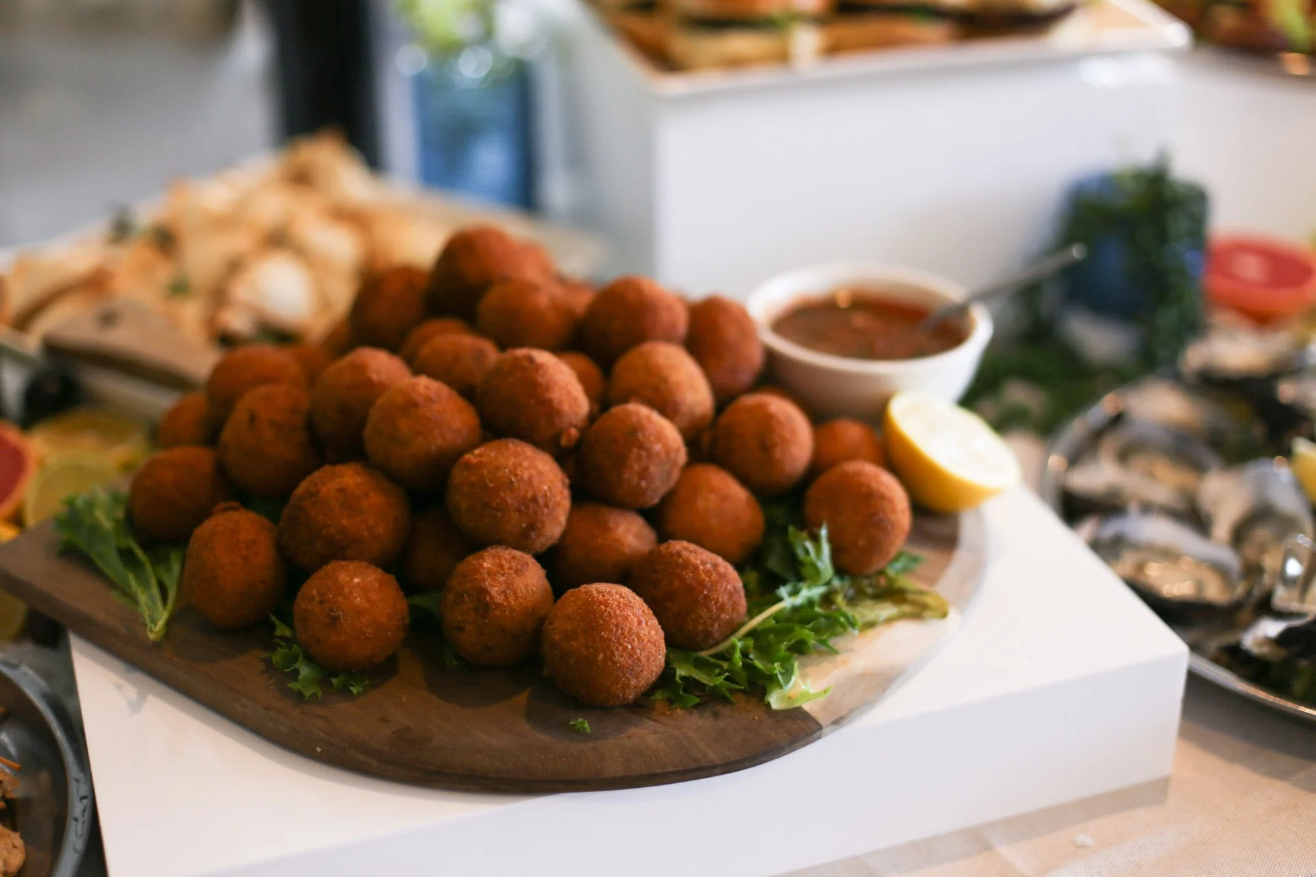 A wooden board full of golden-fried arancini balls with a lemon wedge and green garnishes, with dipping sauce and other dishes blurred in the background.