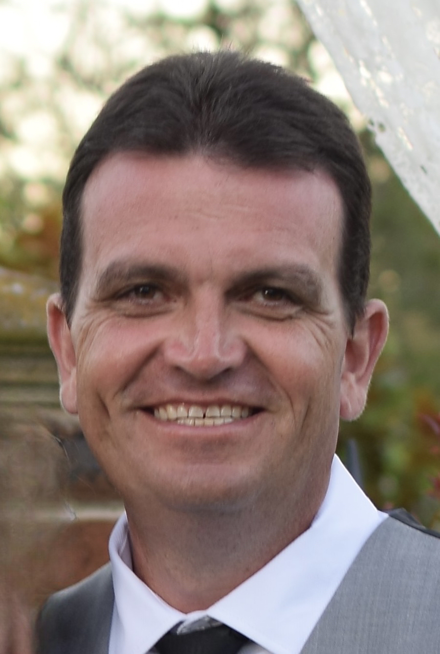 A close-up of a smiling man with dark hair, wearing a light gray suit, white shirt, and black tie, standing outdoors with blurred greenery in the background.