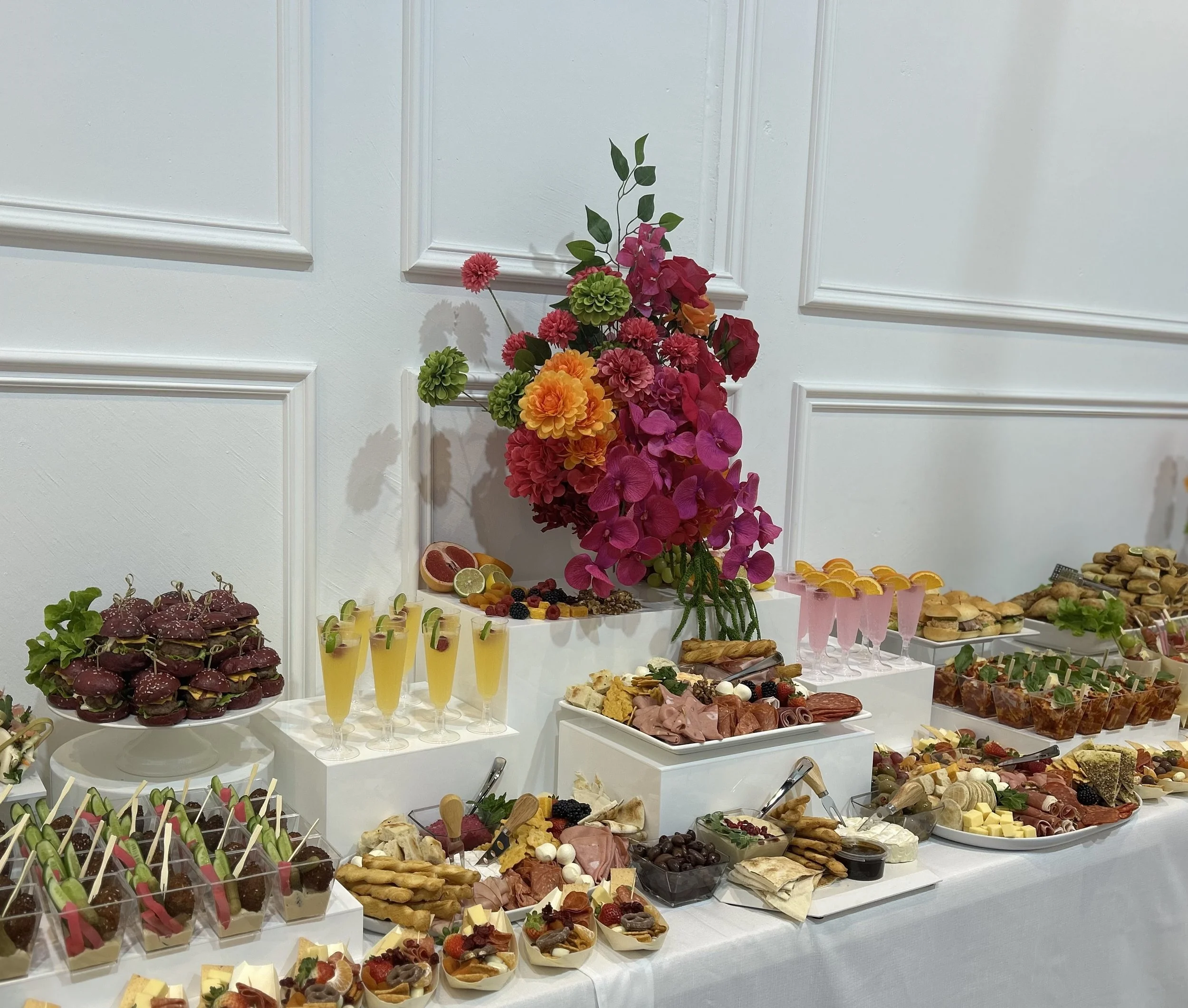 A buffet table with assorted appetizers, desserts, drinks, and a floral centerpiece in a white room.
