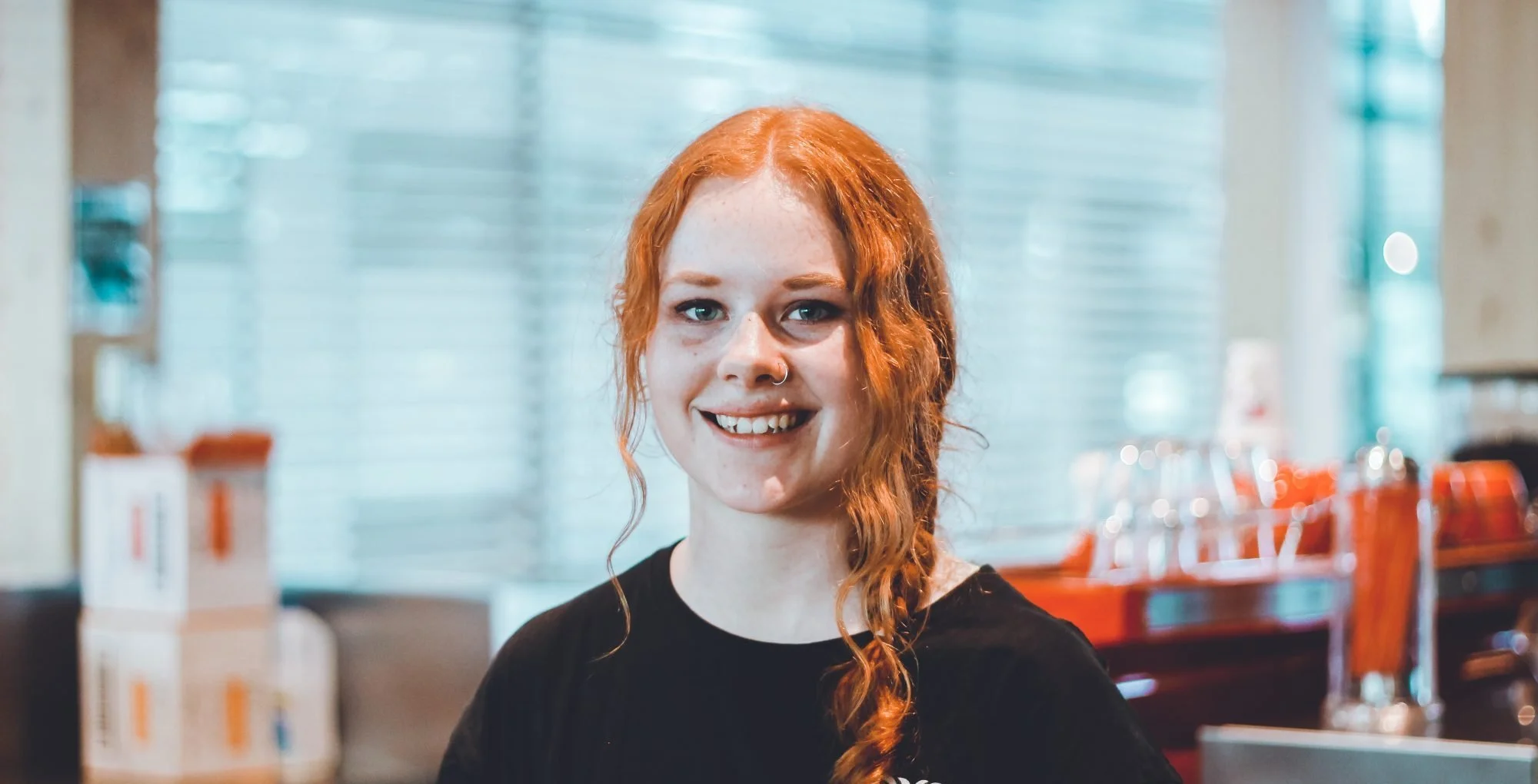 A young woman with curly red hair, pale skin, and a nose ring, smiling in a restaurant or cafe with natural light and wooden decor.