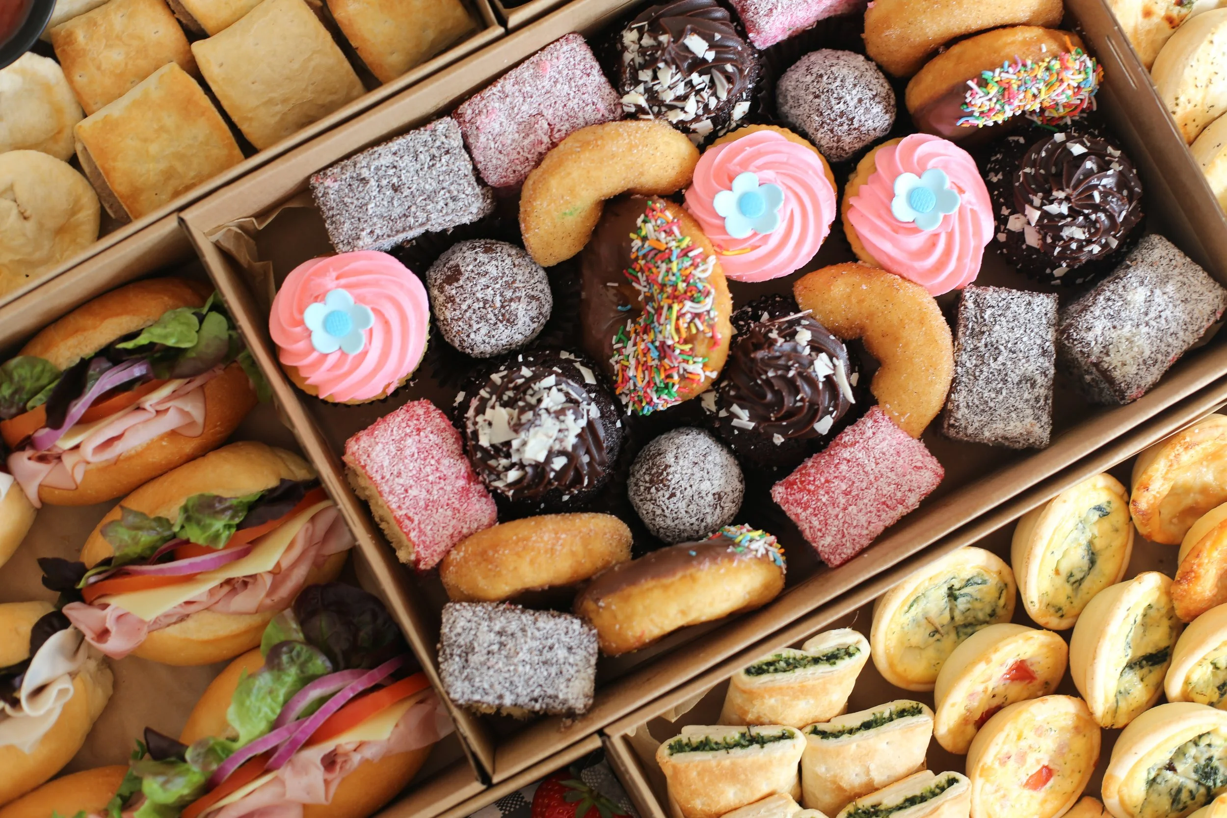 Variety of baked goods including cupcakes with pink frosting and blue flower toppers, chocolate-covered treats with sprinkles, fruit-filled pastries, and mini sandwiches with ham and vegetables.