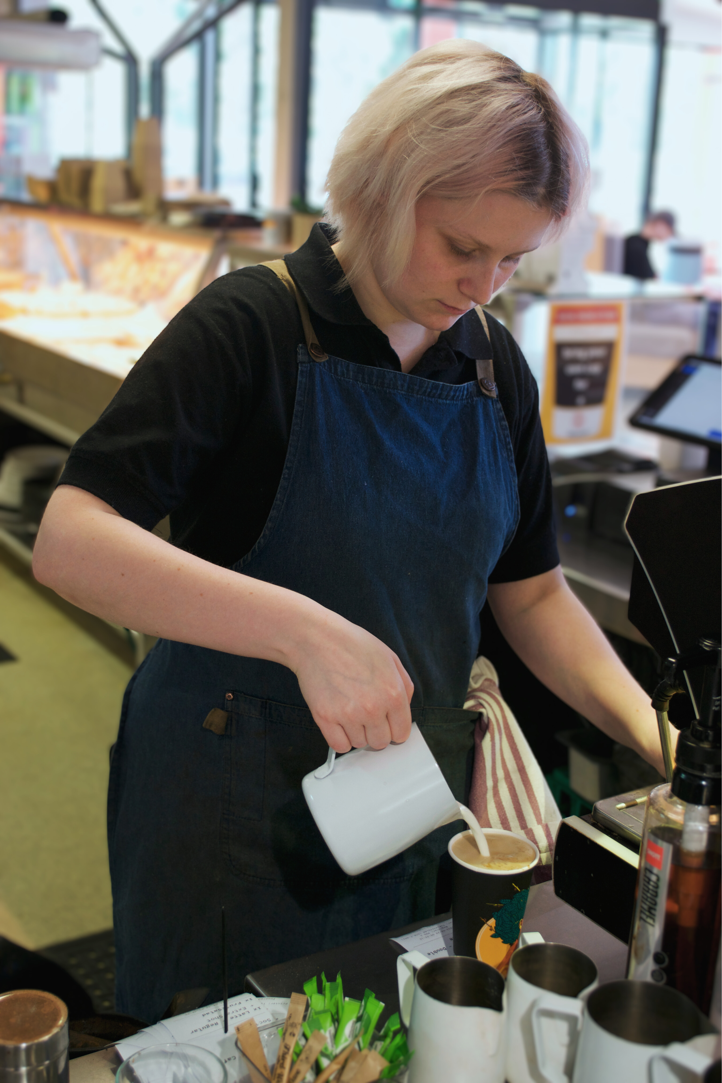 A woman with short blonde hair wearing a black shirt and denim apron is pouring milk into a coffee cup at a counter in a coffee shop.
