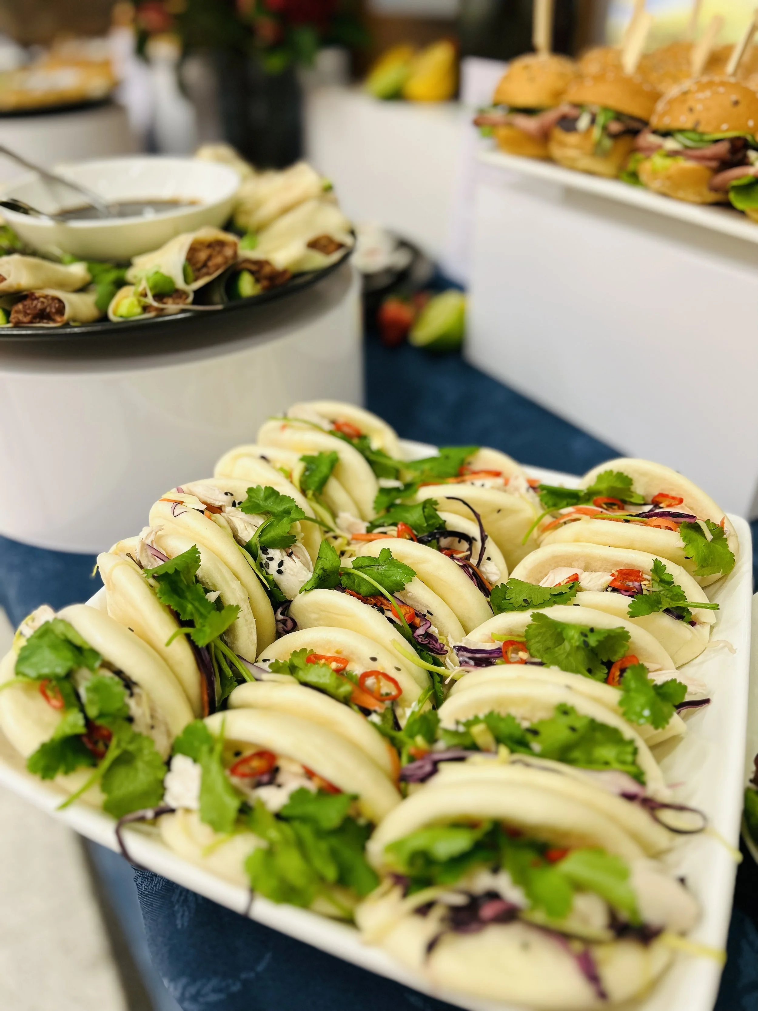 Plate of steamed bao buns filled with pork and garnished with cilantro, red chili slices, and purple cabbage, along with other sandwiches and appetizers in the background.
