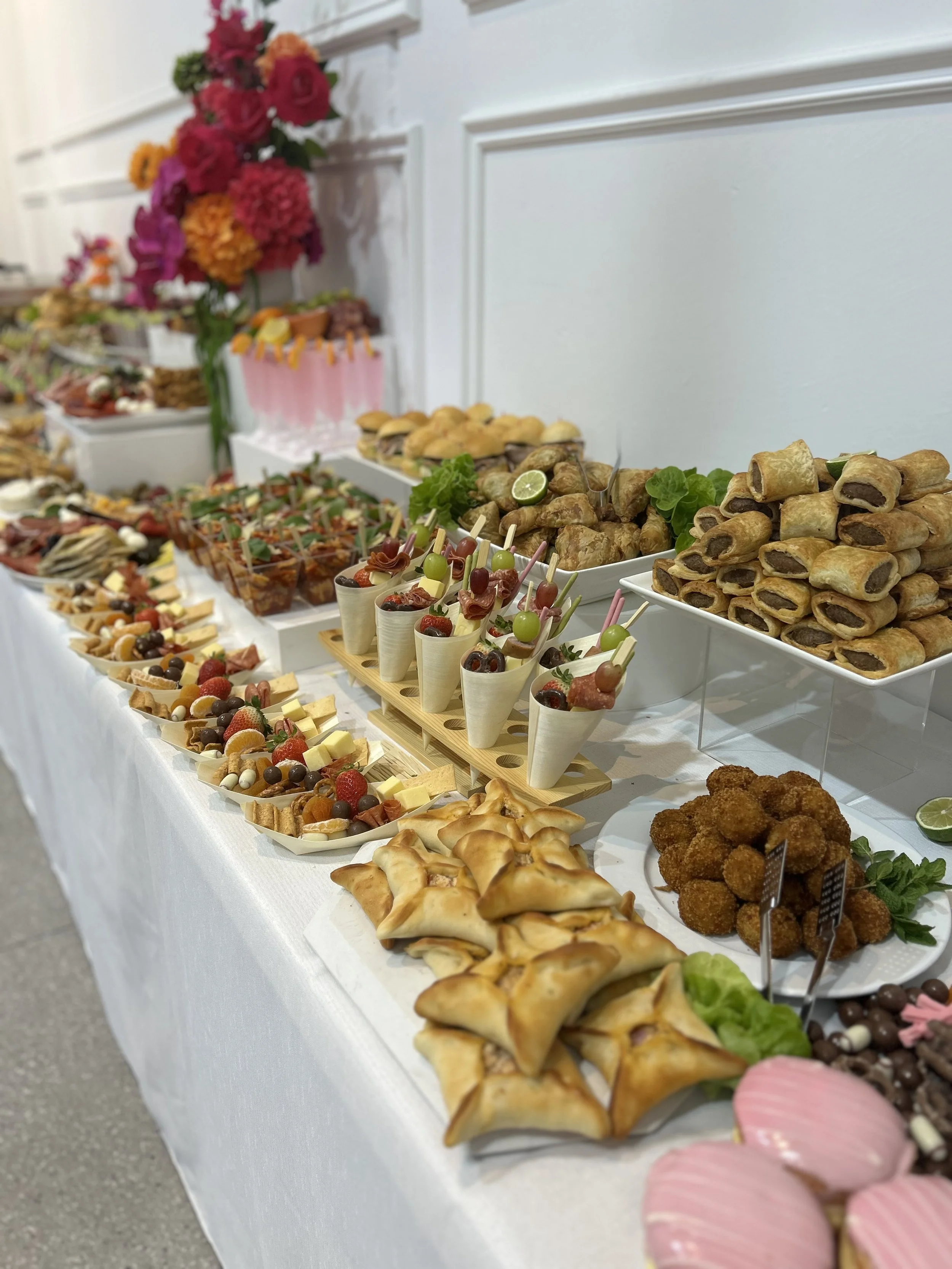 A buffet table with various appetizers and finger foods, including mini sandwiches, spring rolls, fruit and cheese platters, skewers, and fried bites, decorated with lime slices and green herbs.