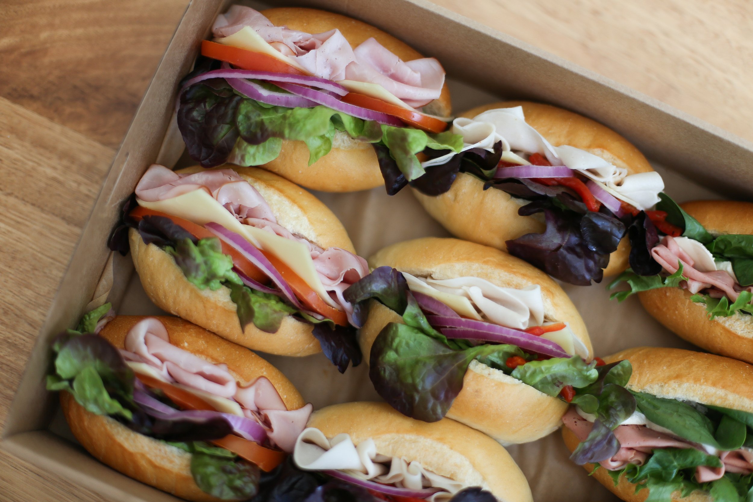 A tray of mini sandwiches with deli meats, cheese, fresh lettuce, tomato, purple onion, and sliced red pepper on small French rolls.