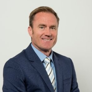 Portrait of a man in a suit with a blue shirt and striped tie, smiling against a plain background.